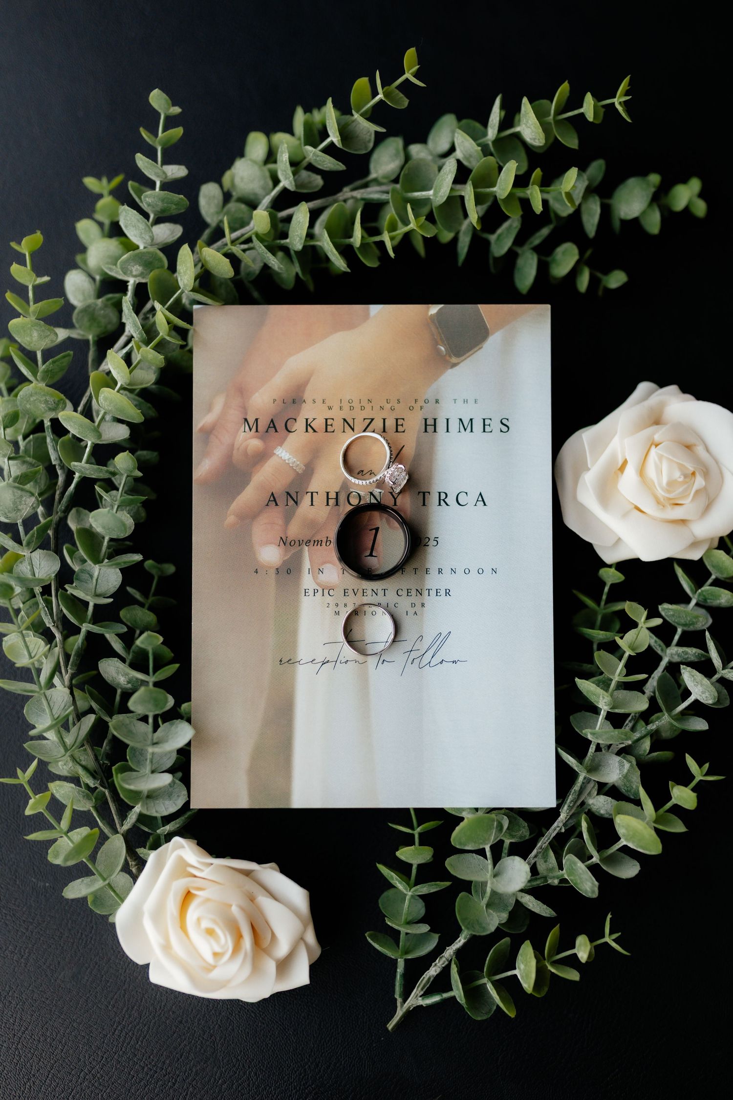 Wedding invitation for Mackenzie Himes and Anthony Trca with rings, roses, and eucalyptus on black.