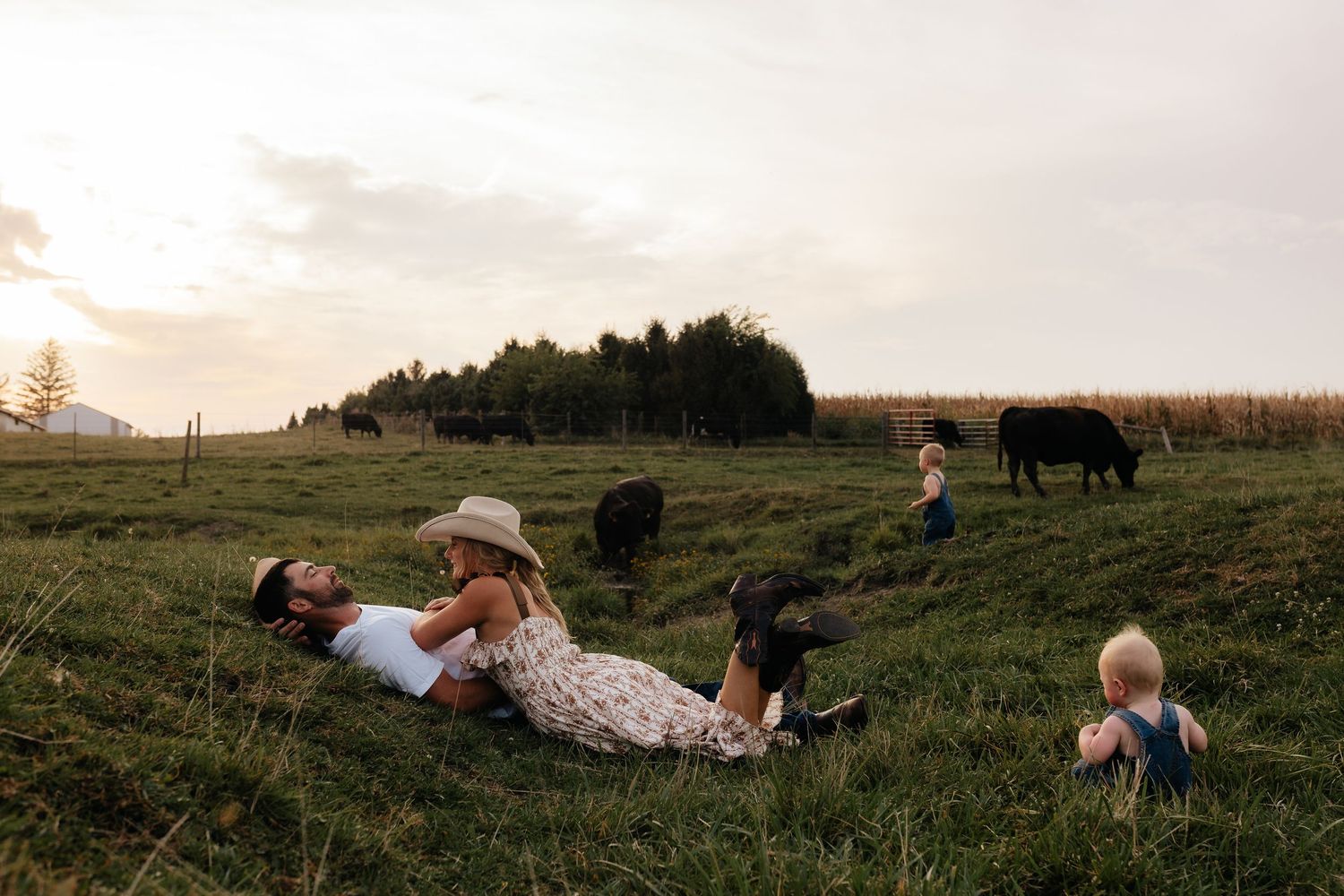 Family relaxing in farm field at sunset with cattle grazing in background on rural property.
