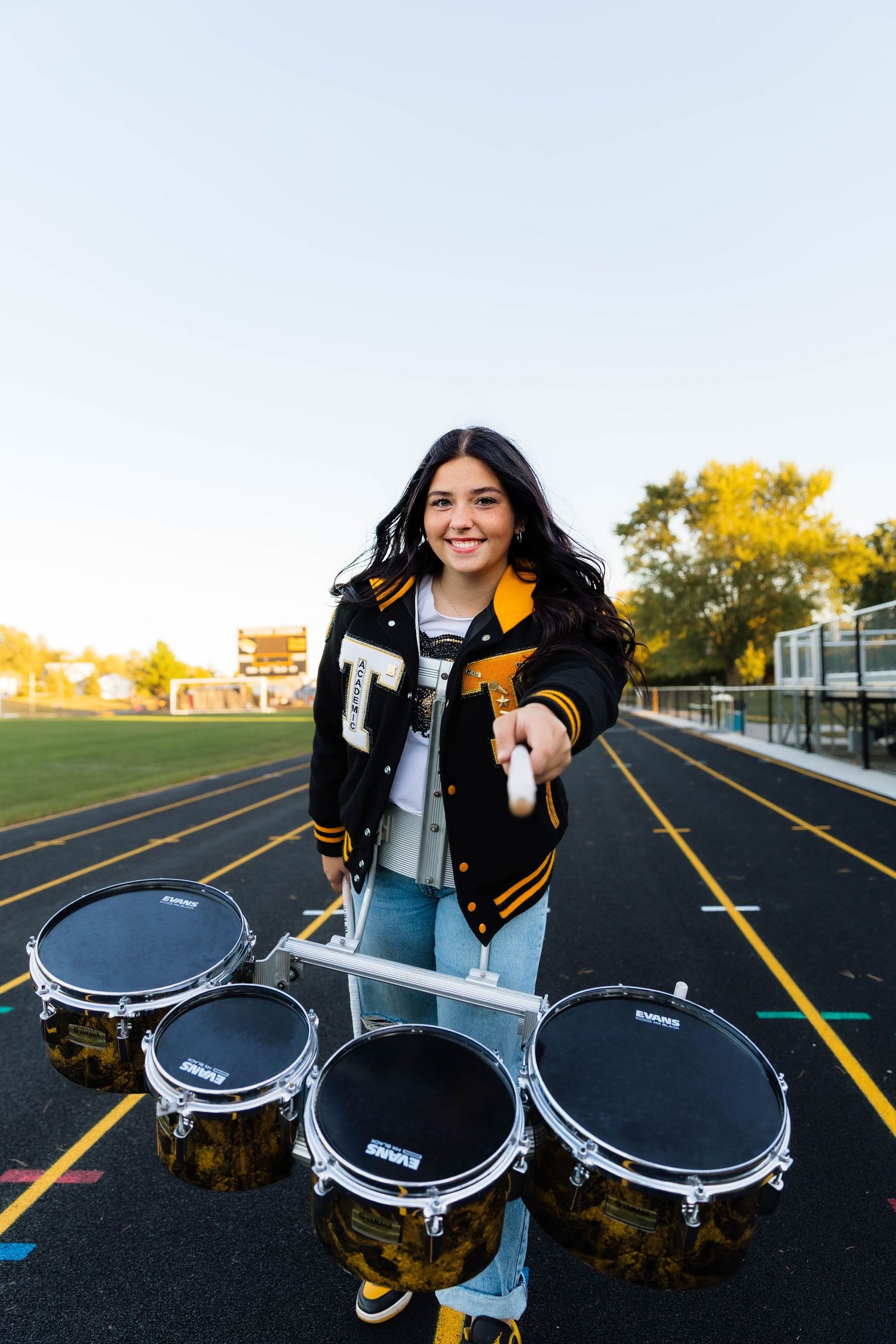Smiling teen in varsity jacket poses with tenor drums on high school track at golden hour.