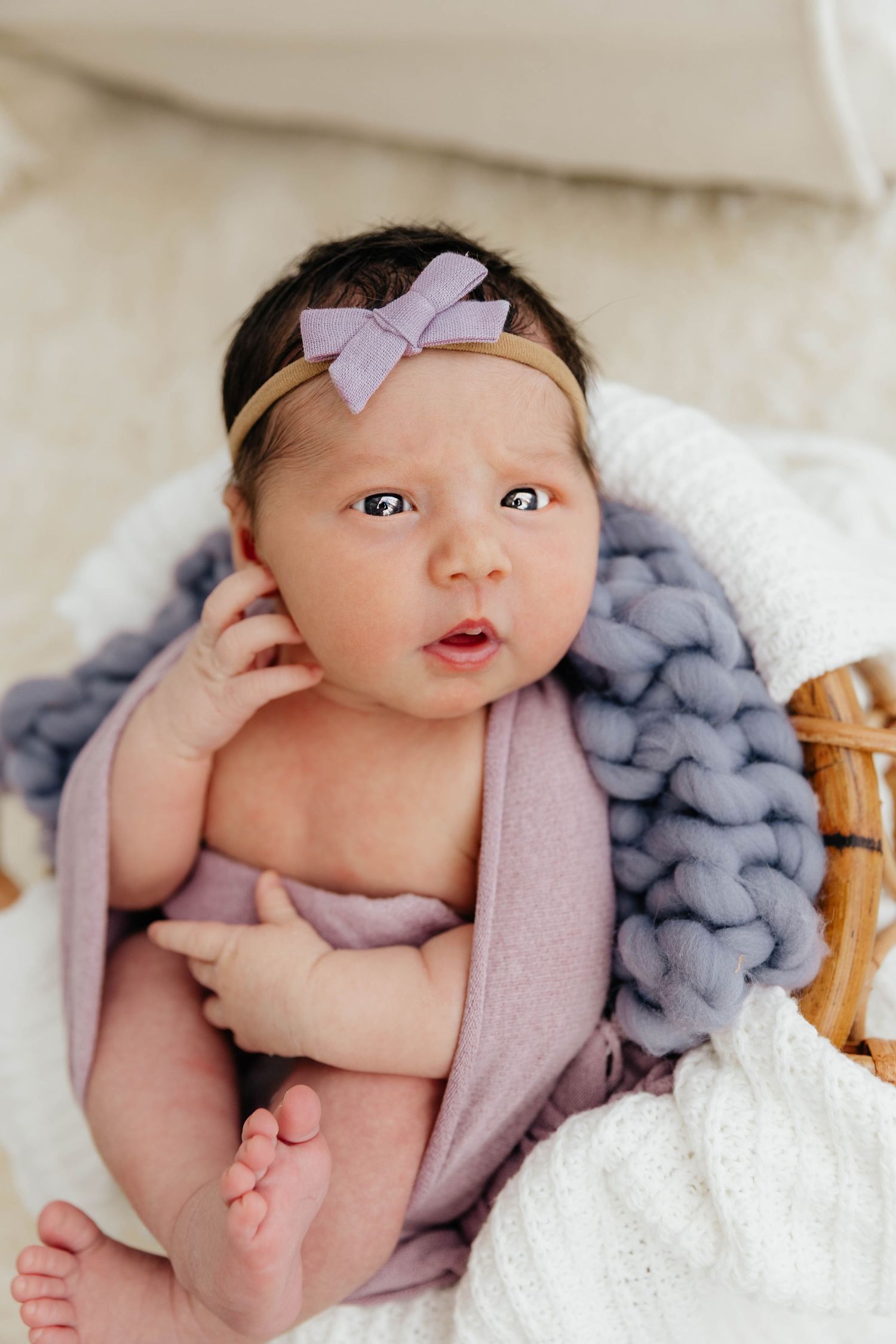 Wide-eyed newborn baby in lavender wrap and purple bow headband sitting in wicker basket.