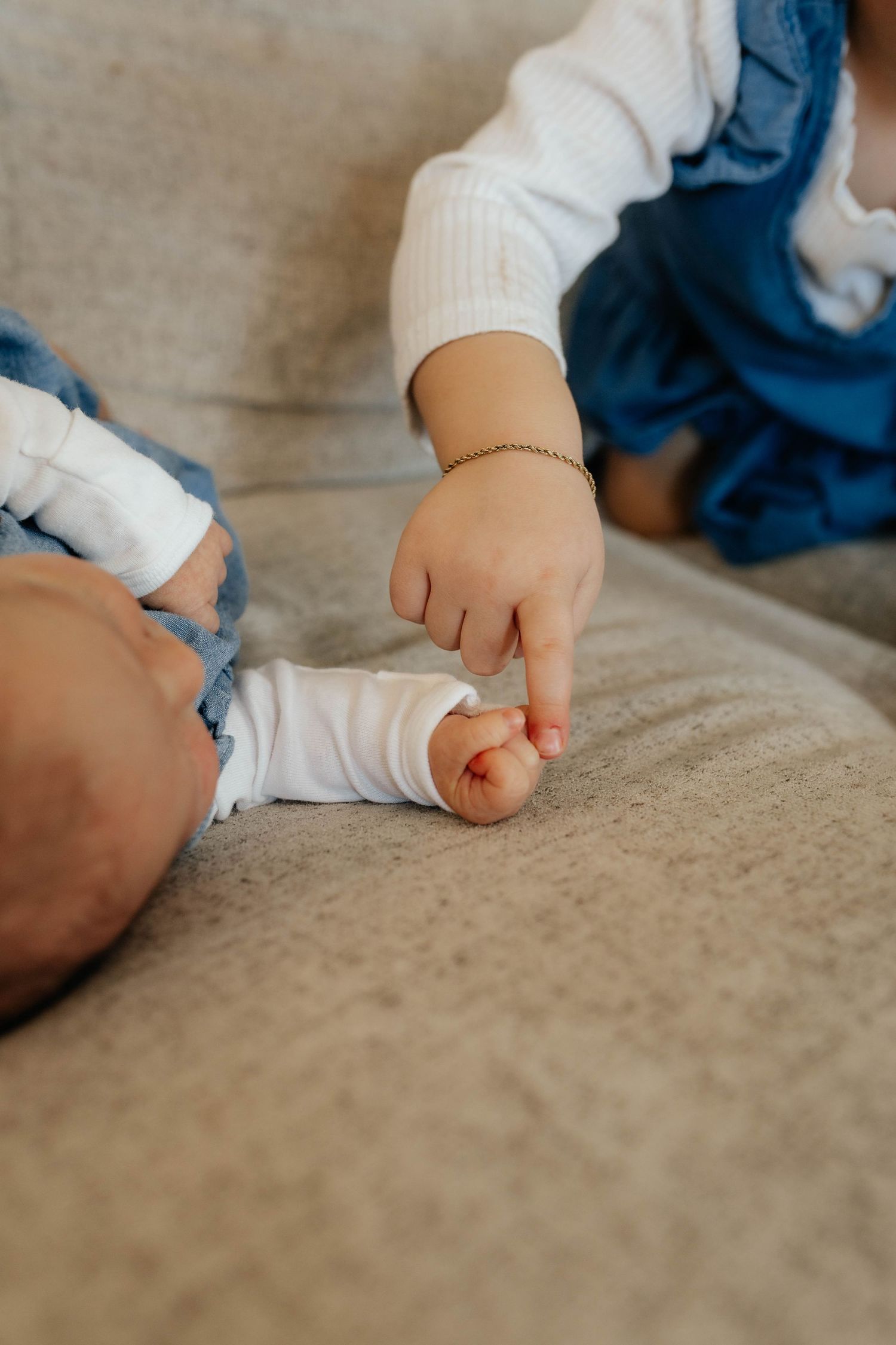 Close-up of a toddler gently touching a newborn baby's hand on a beige sofa.