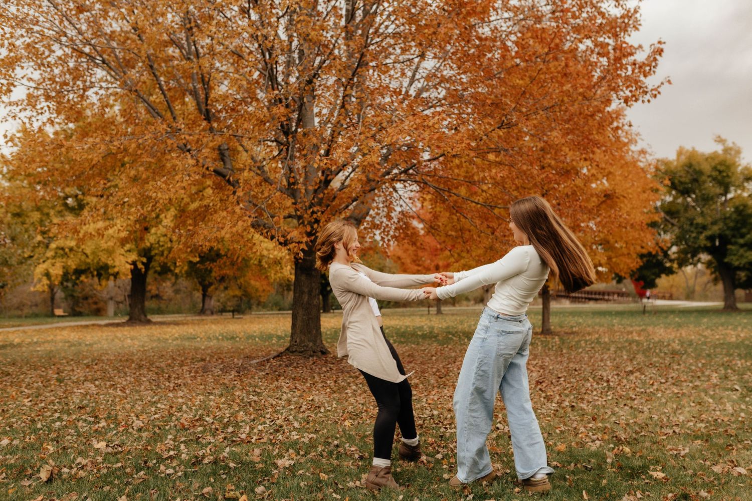 Two teenage girls spinning and holding hands together in front of vibrant orange autumn foliage.