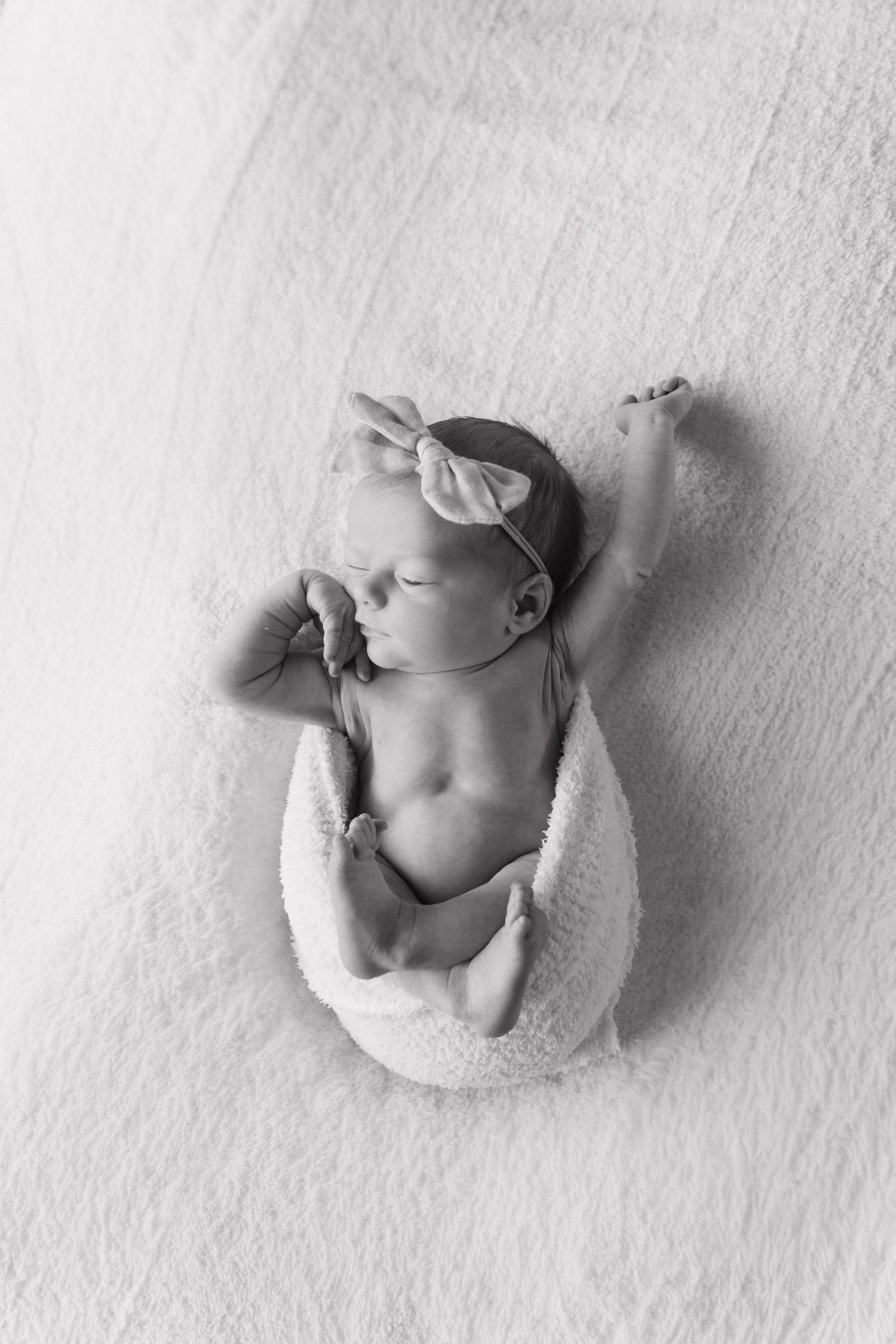 Black and white photo of sleeping newborn baby girl with bow headband on white blanket.