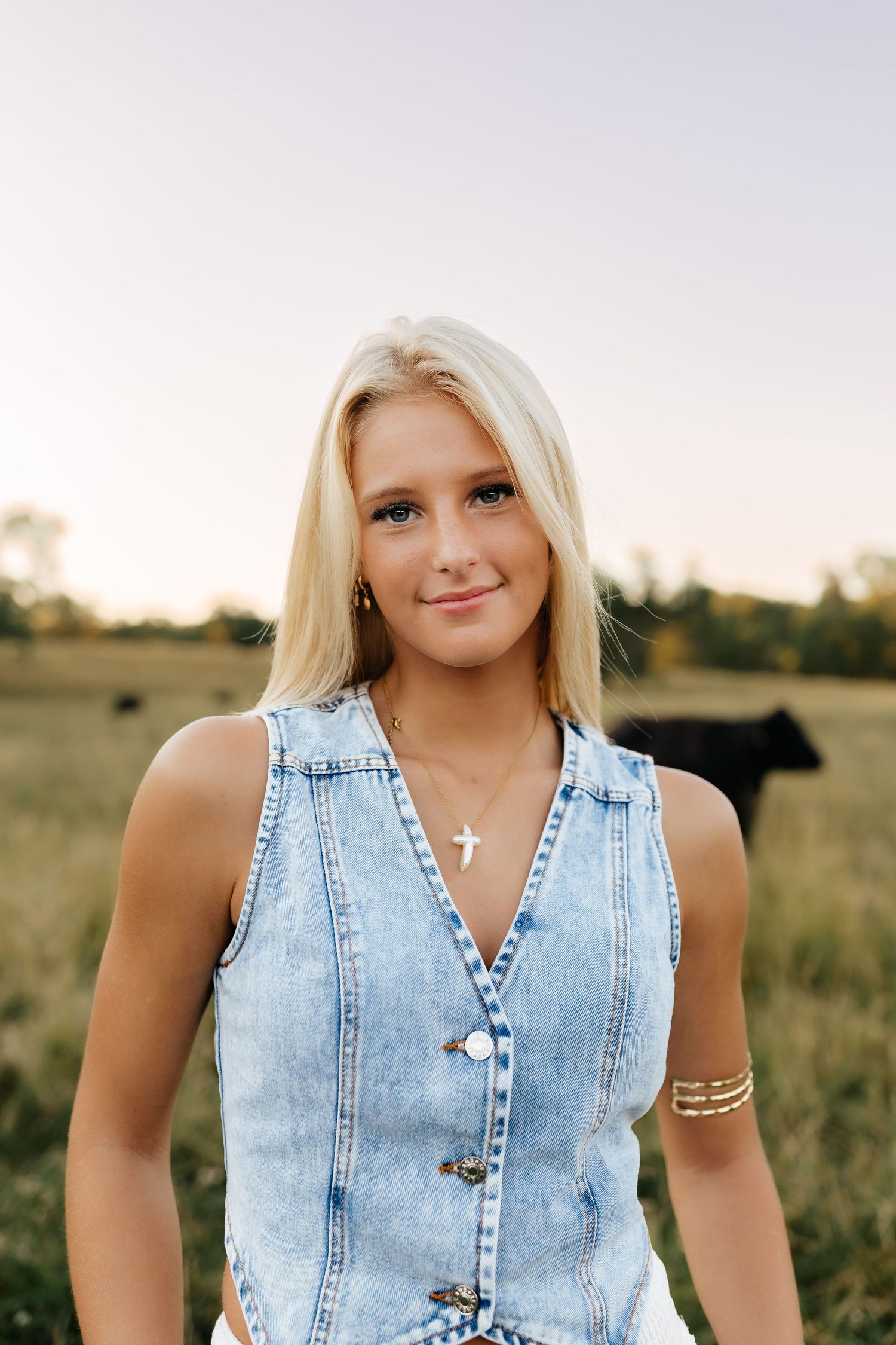 Blonde teenage girl in denim vest with cross necklace smiling in a field with cattle at sunset.