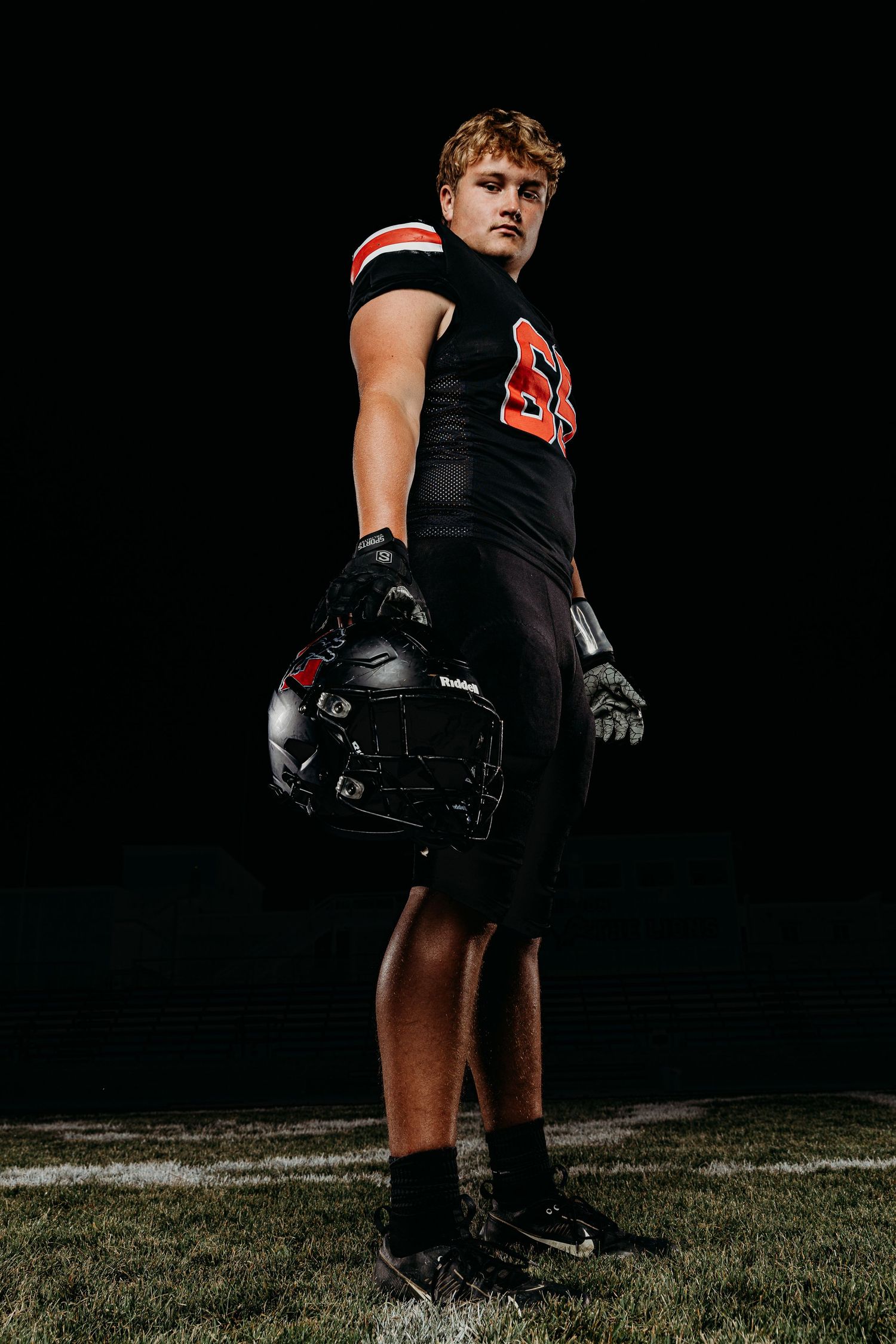 High school football player in black uniform holding helmet under stadium lights on dark field.