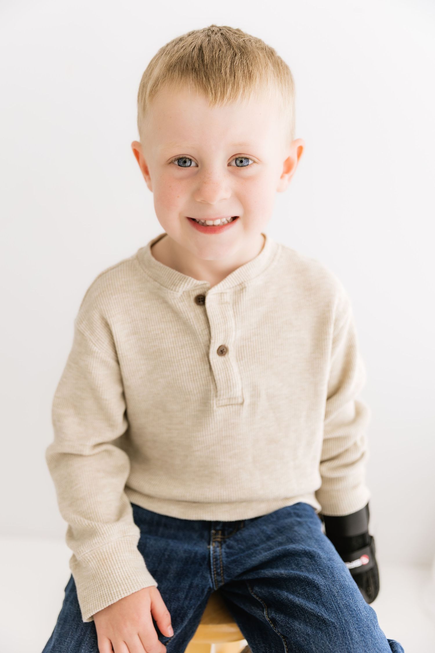 Smiling blonde toddler boy in beige henley shirt sits on wooden stool in bright white studio.
