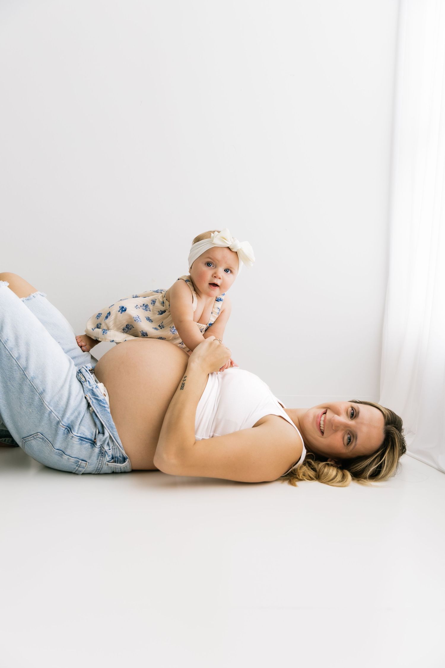 Pregnant woman lying on floor smiling while her baby rests on her bare belly in a bright white room.