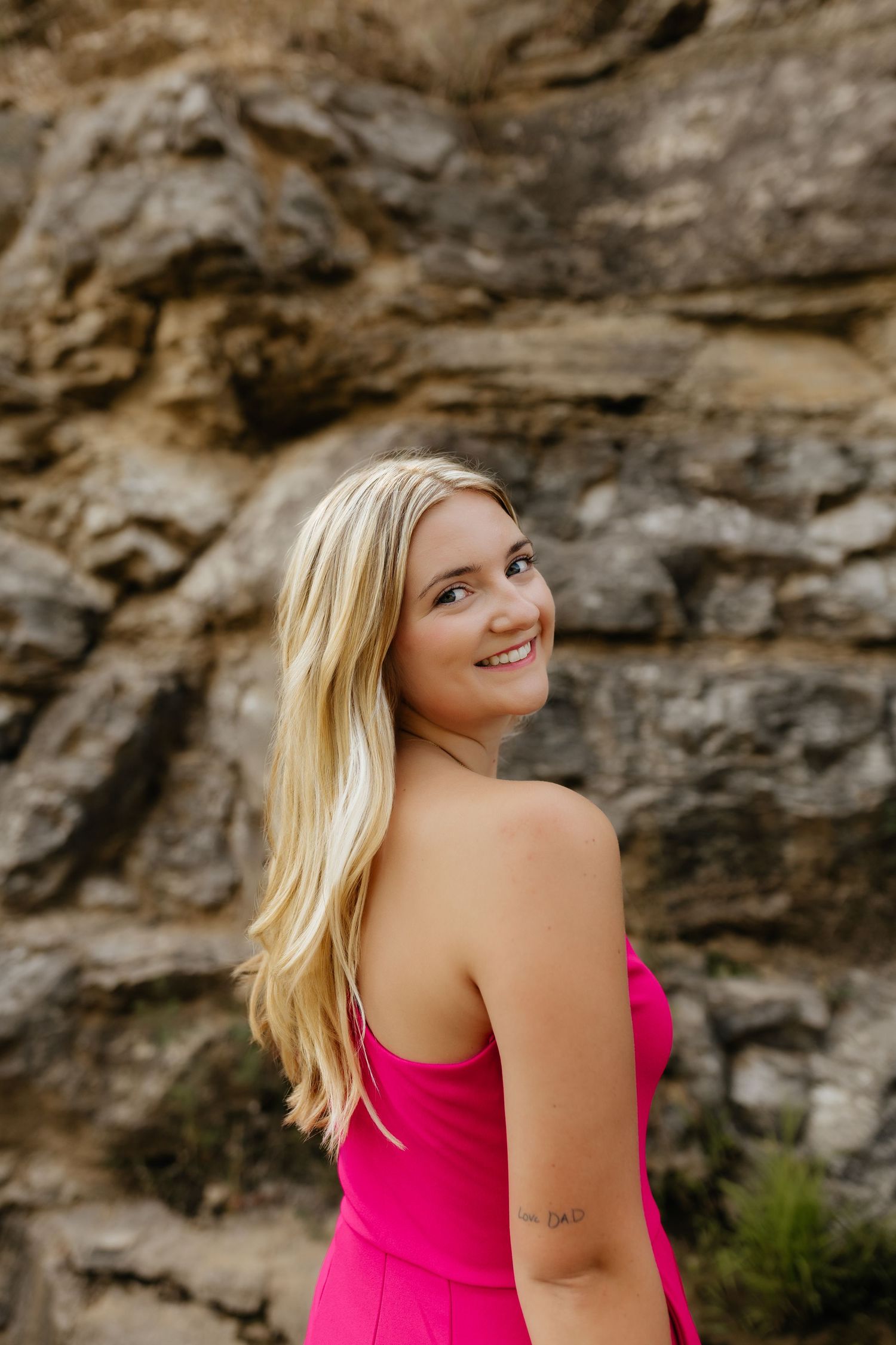 Smiling blonde woman in a hot pink strapless dress posing against a rocky stone wall backdrop.