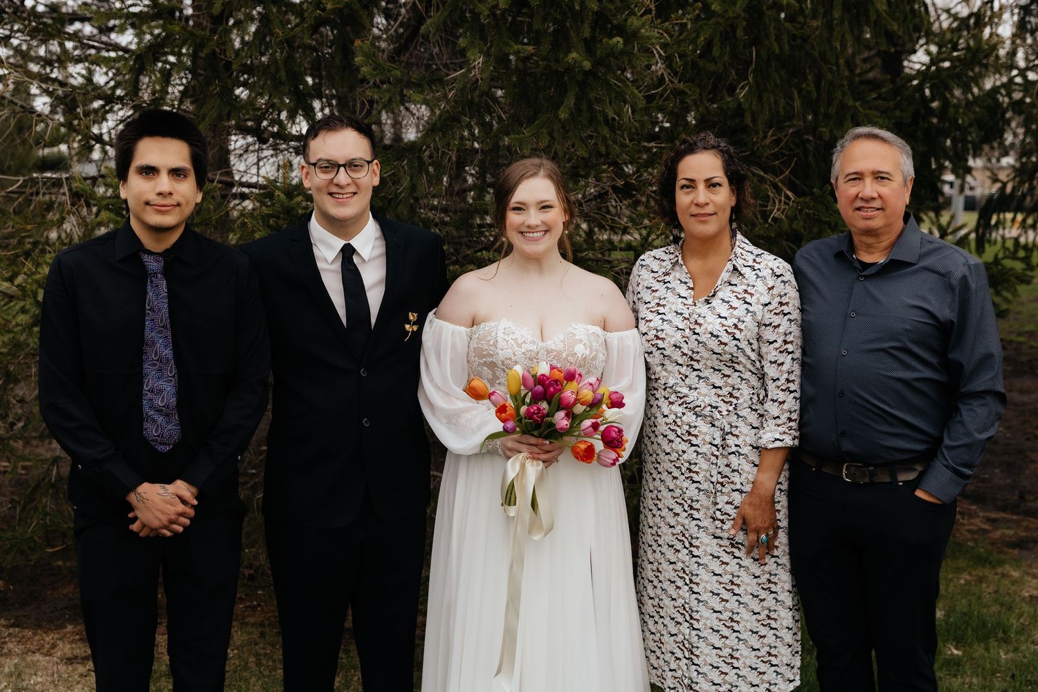 Wedding couple and family of three pose outdoors near trees after ceremony.