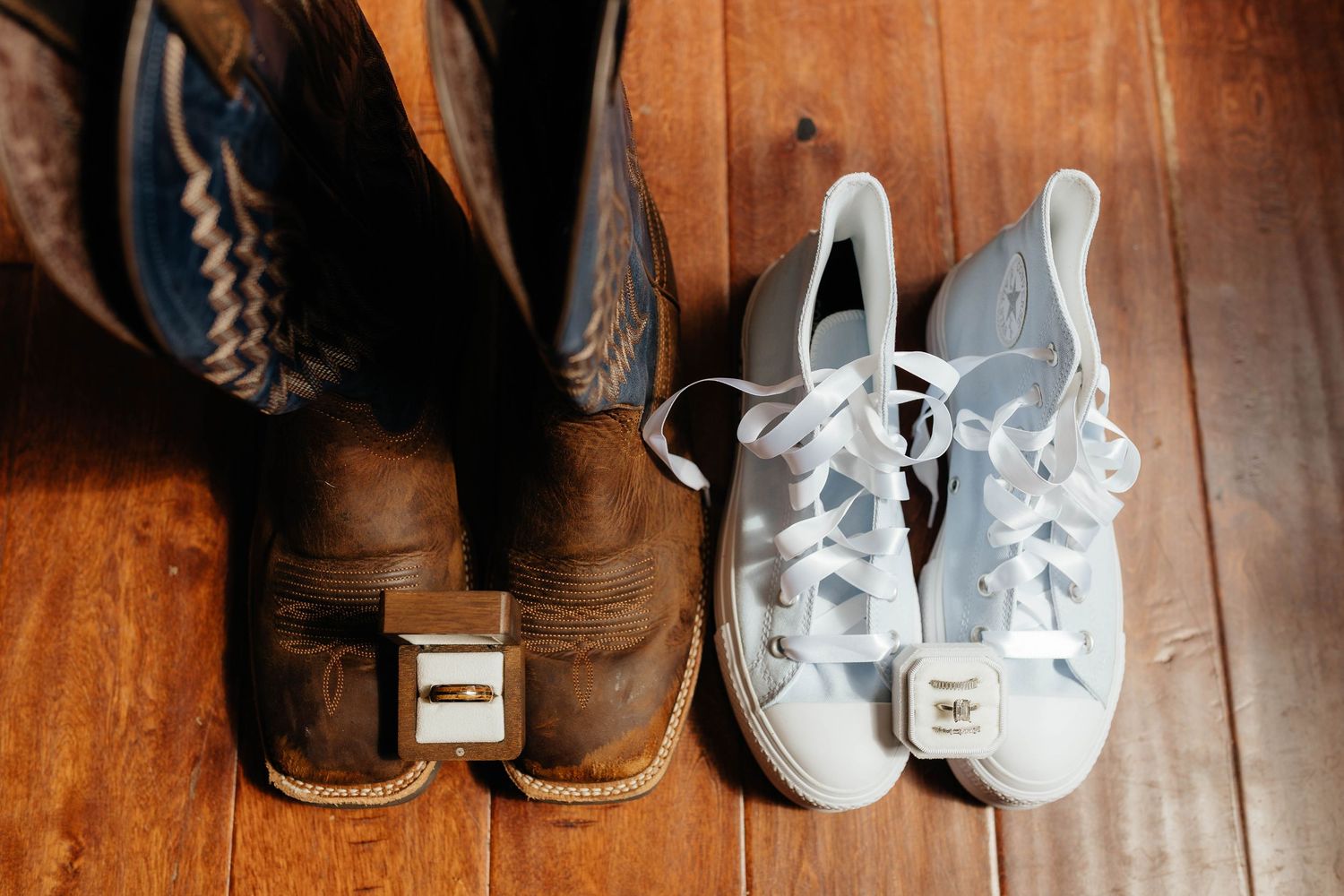 Cowboy boots and white Converse sneakers with wedding rings placed between them on hardwood floor.