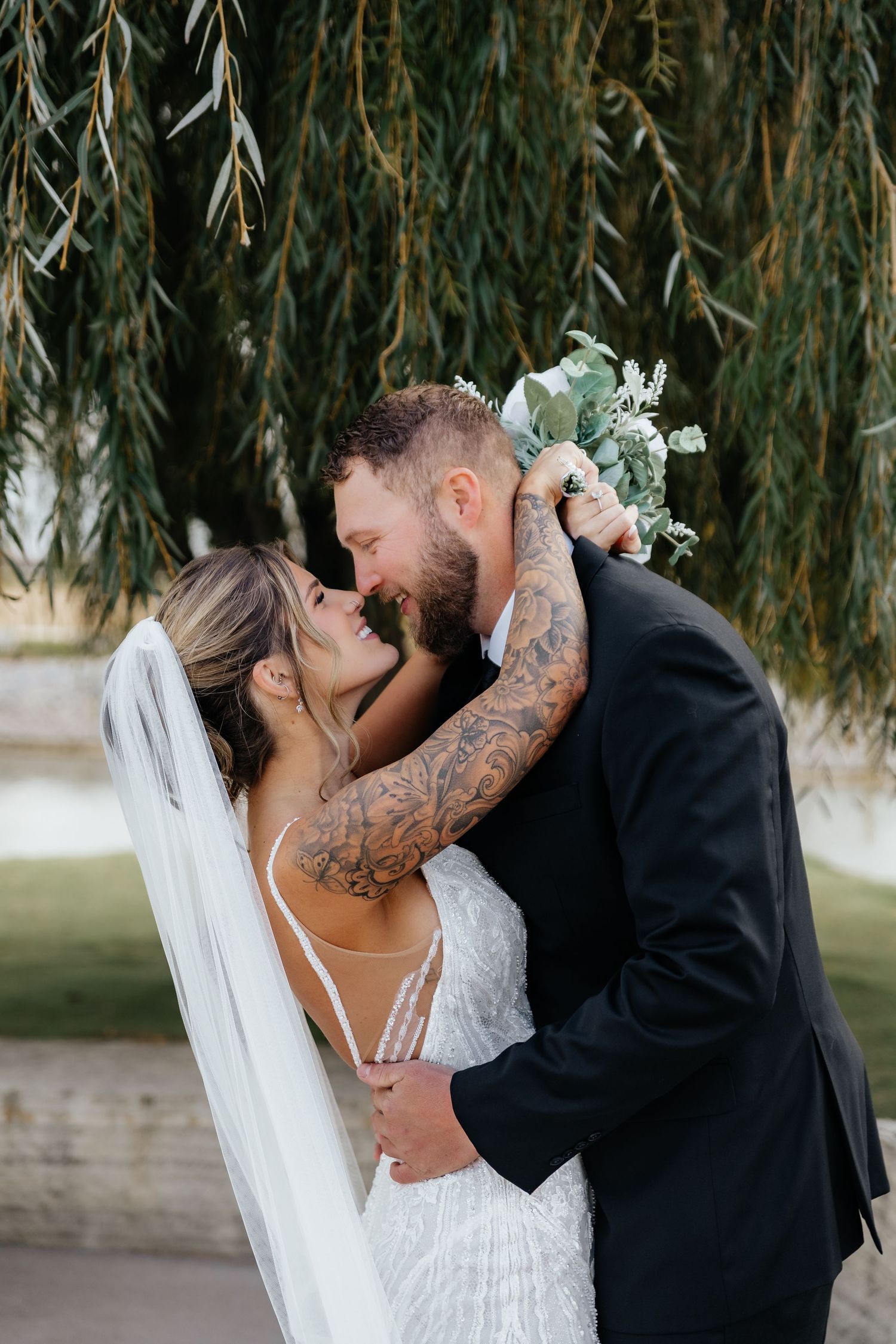 Tattooed groom holding bouquet kisses bride in lace wedding dress under weeping willow tree.