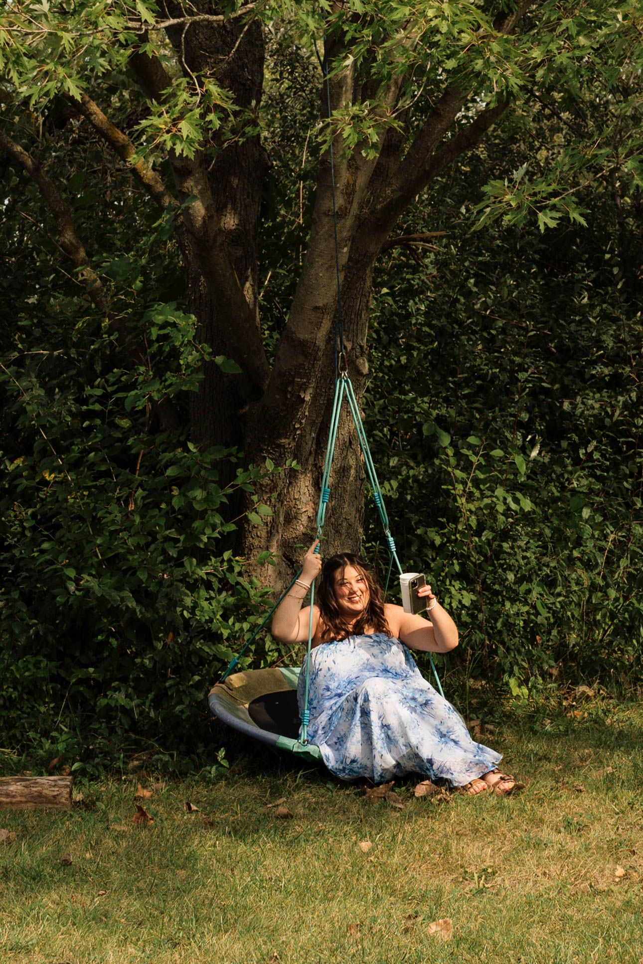 Woman in blue floral dress sits smiling on a circular tree swing in a lush green garden.