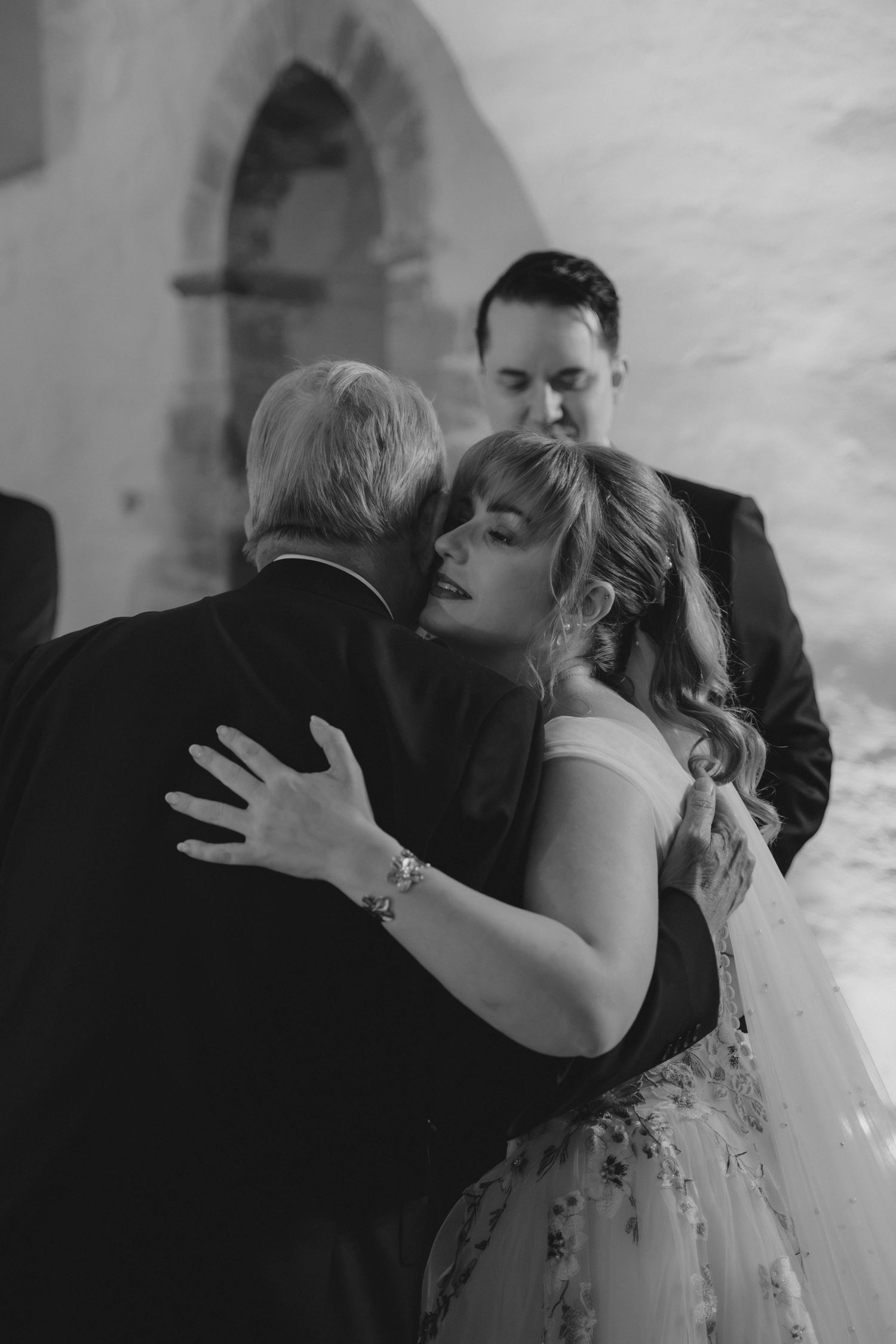 Emotional black and white photo of bride embracing a guest while groom stands nearby at wedding.