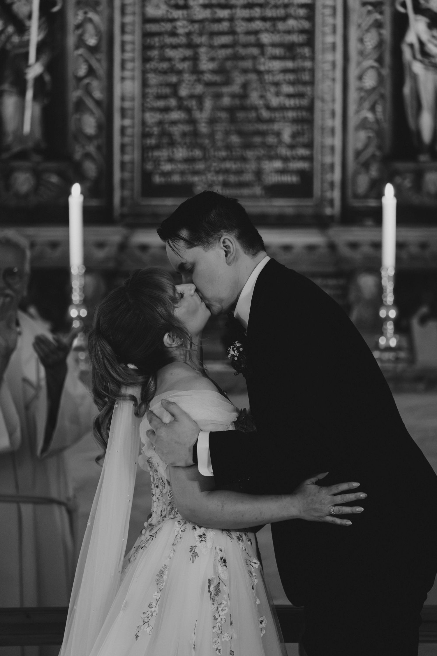 Bride and groom share first kiss at altar with candles and ornate backdrop in black and white.