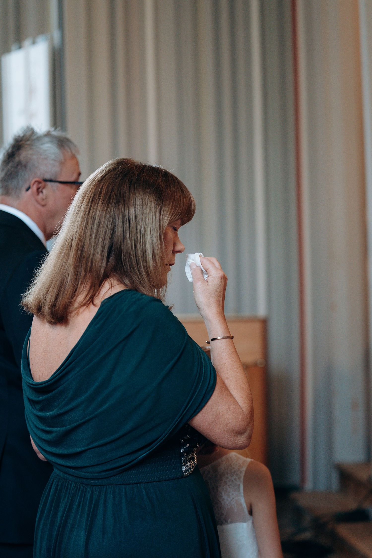 Emotional wedding guest in teal dress dabbing tears with tissue during ceremony.