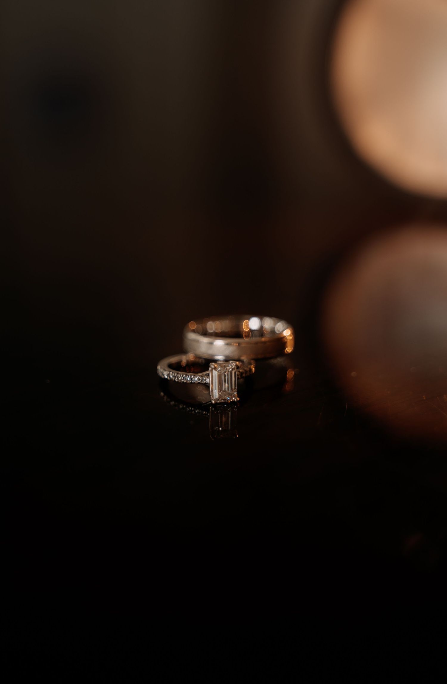 Three stacked wedding rings with diamonds on a dark reflective surface, featuring an emerald-cut engagement ring.