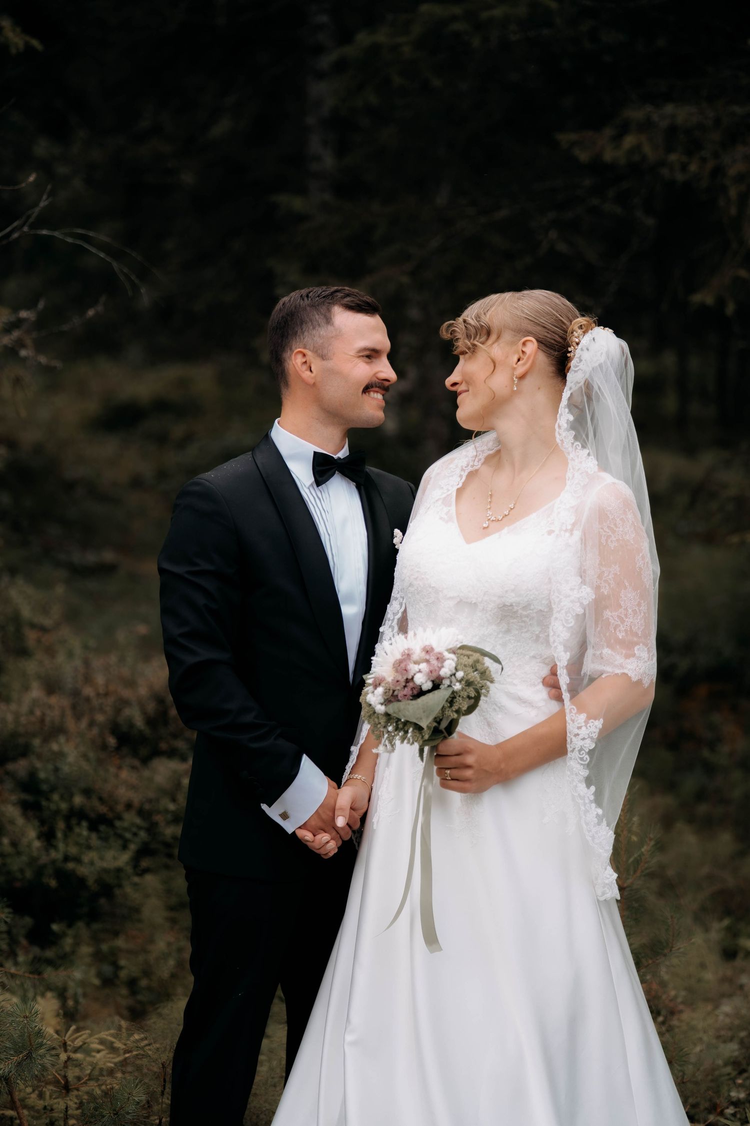 Bride and groom holding hands smiling at each other in a forest, bride in lace gown with veil holding bouquet.