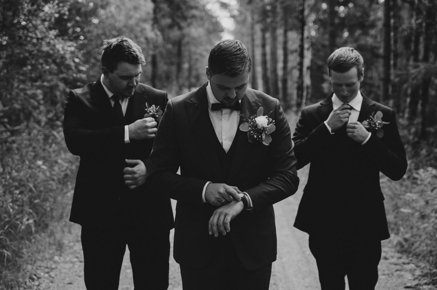 Black and white photo of groom checking watch flanked by two groomsmen adjusting suits on a forest path.