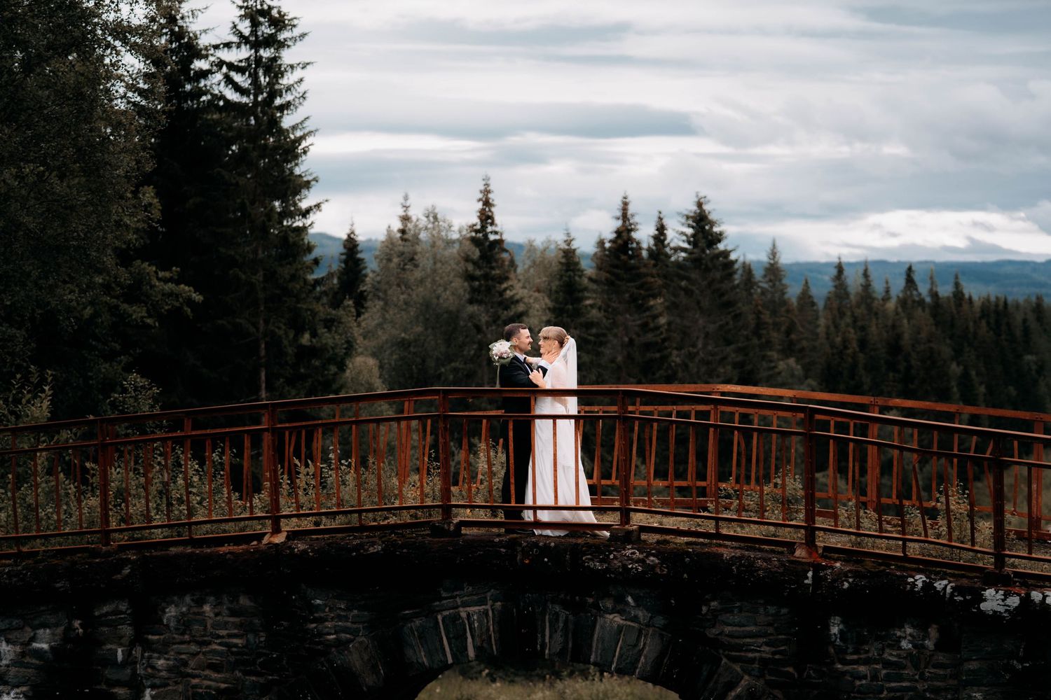 Couple embracing on a rustic wooden bridge surrounded by evergreen forest and mountain views on their wedding day.