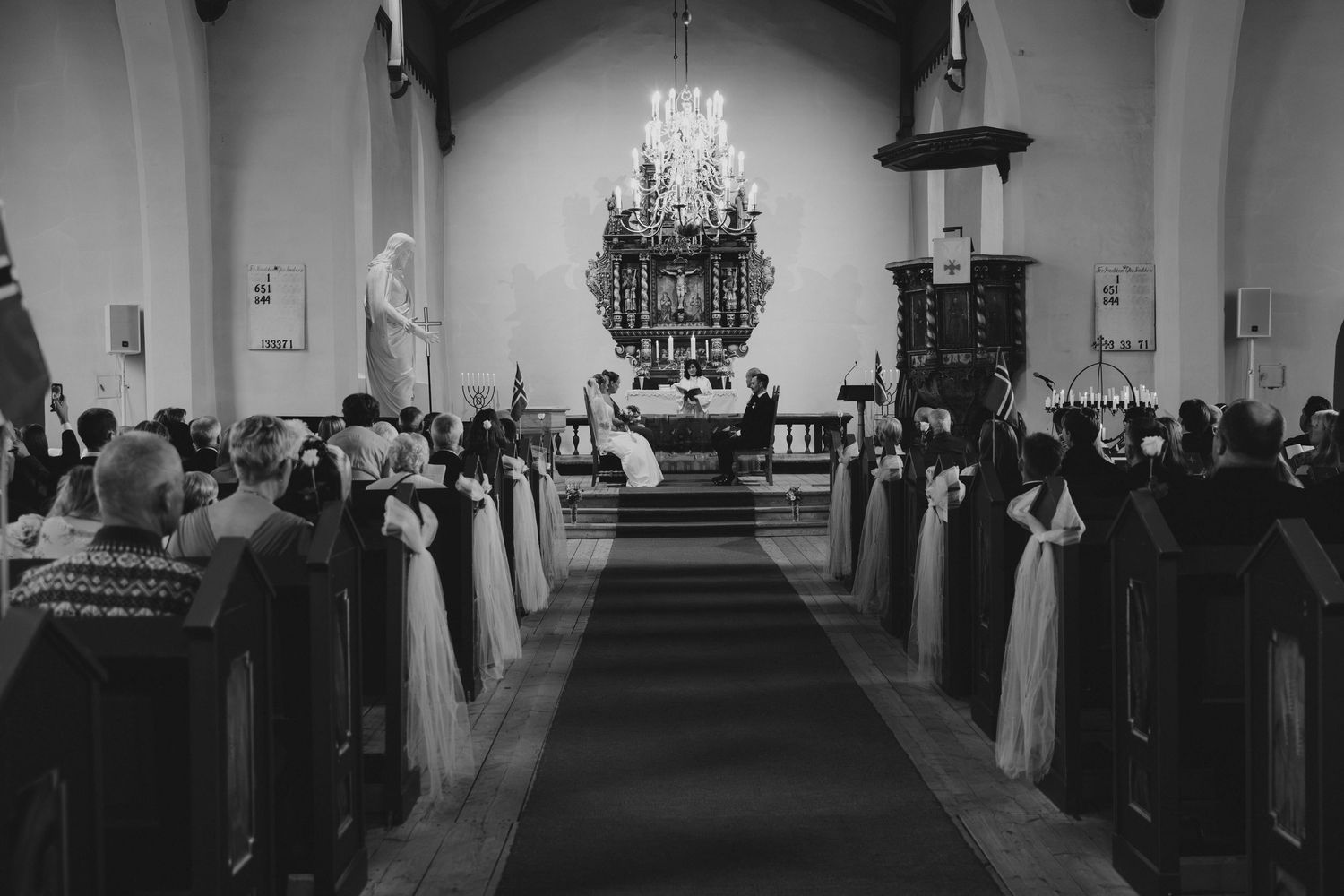 Black and white photo of wedding ceremony inside a church with guests seated in pews and chandelier overhead.