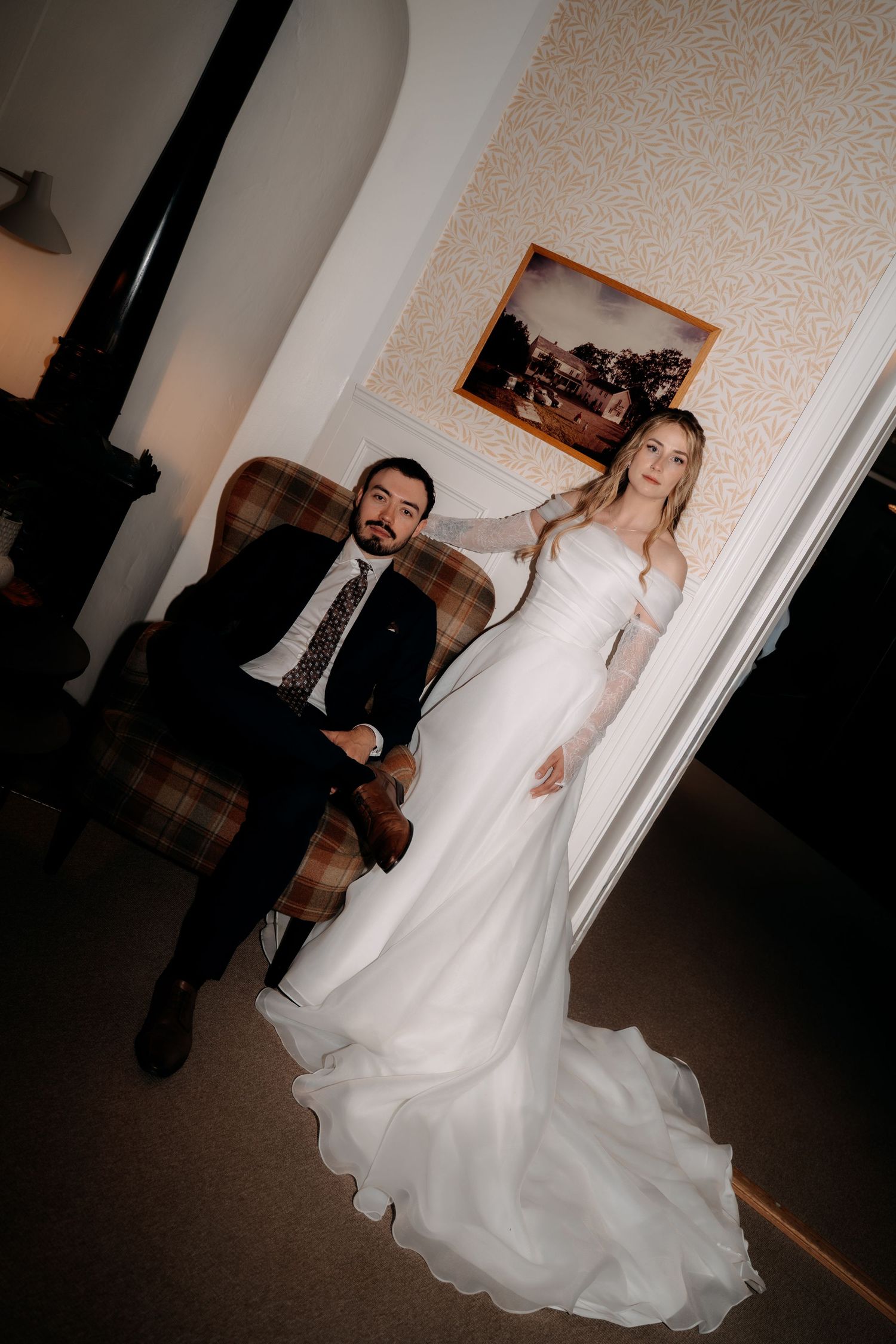 Bride in elegant white gown stands beside groom in dark suit seated in plaid chair during indoor wedding portrait.
