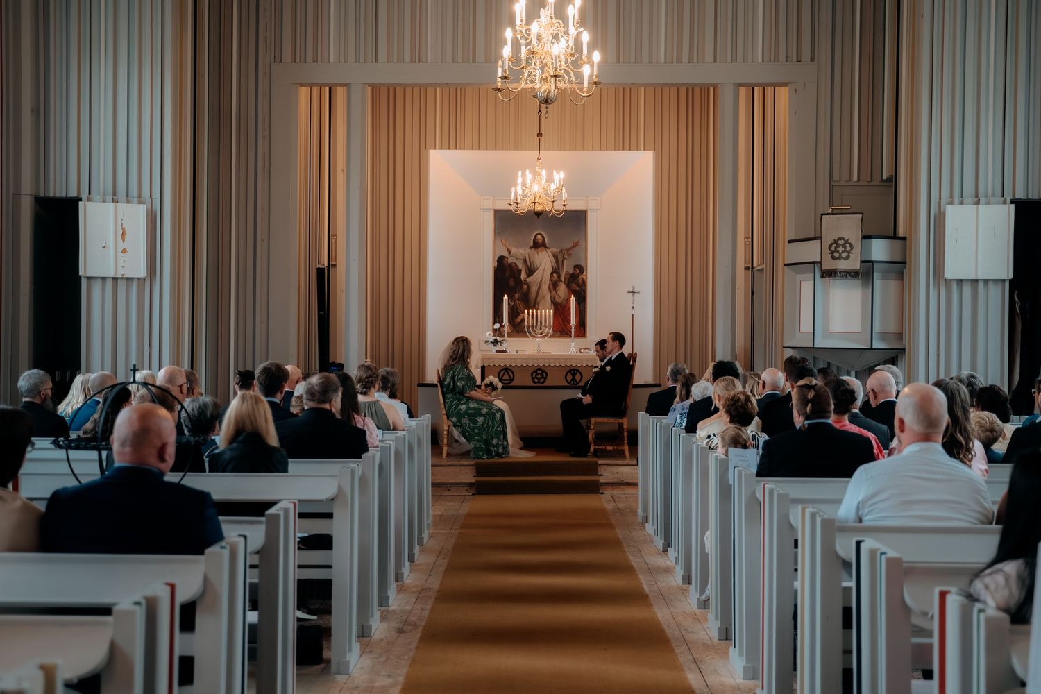 Guests seated in white church pews during an elegant wedding ceremony with chandelier and altar backdrop.