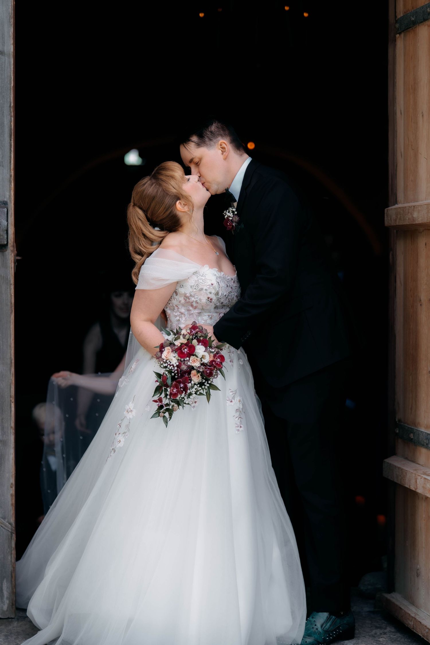 Bride and groom sharing a romantic kiss in a barn doorway, bride holding a red and white bouquet.