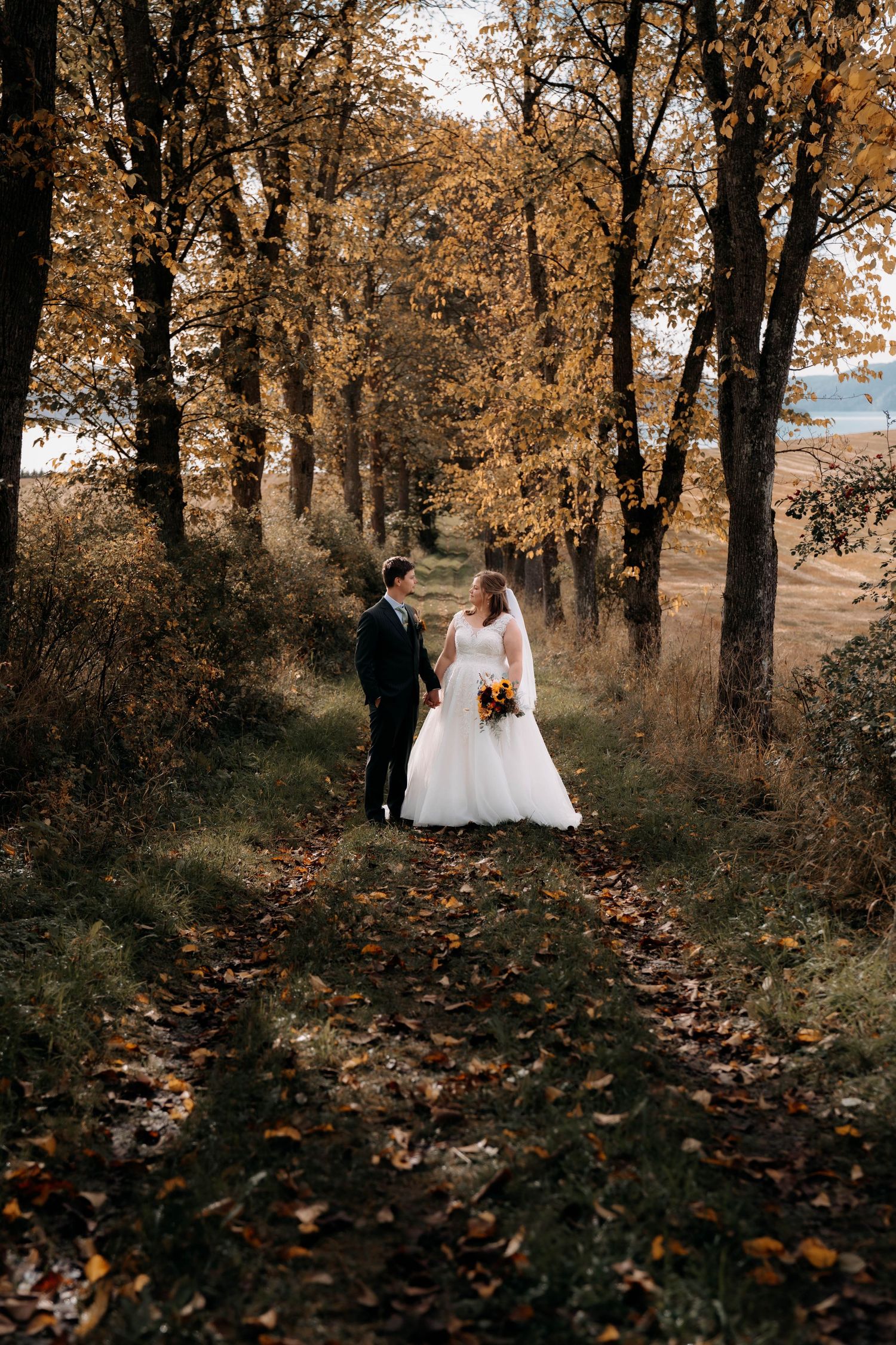 Bride and groom walk hand in hand along a leaf-covered autumn path surrounded by golden trees.