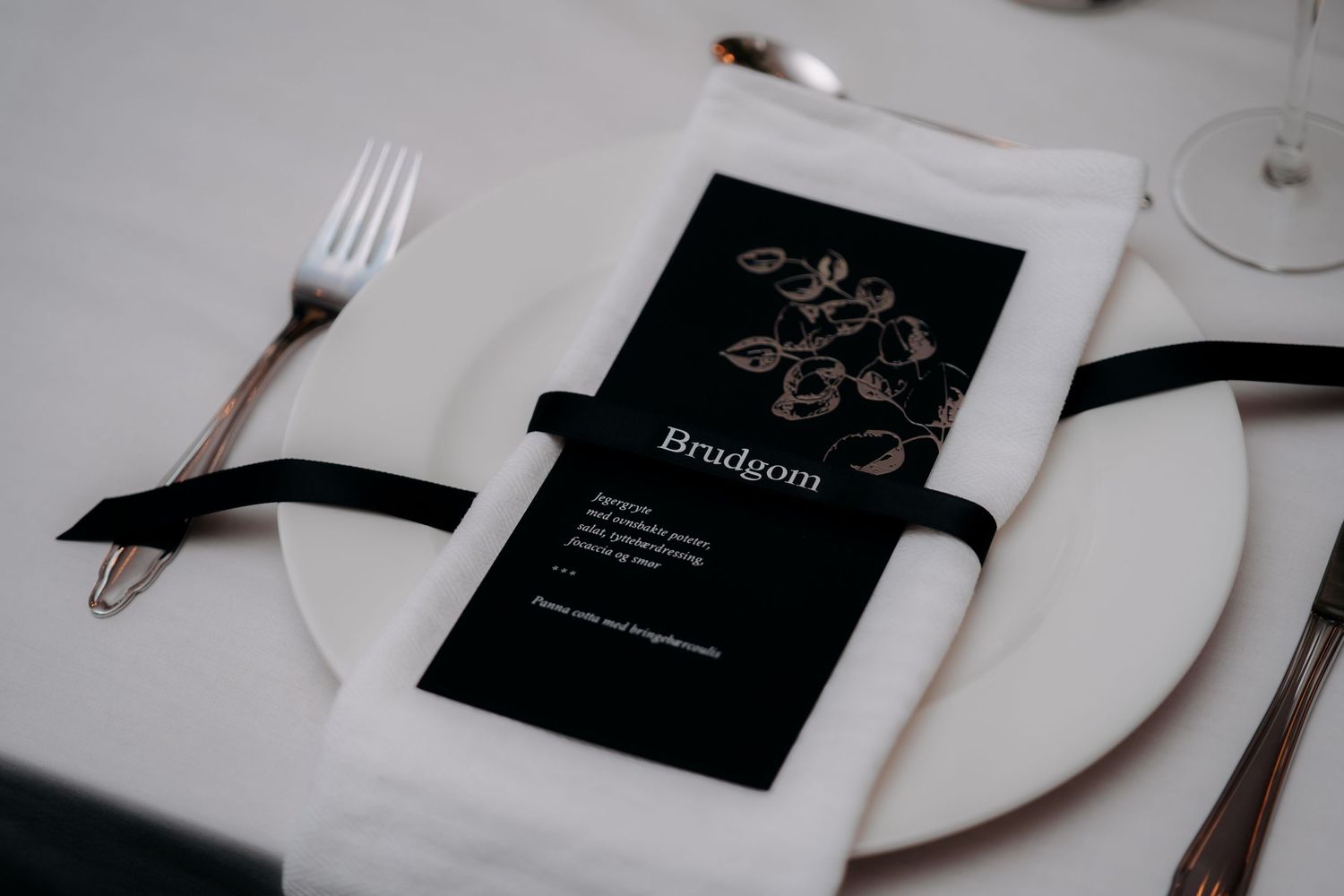 Elegant wedding place setting with black menu card reading Brudgom, white napkin, and rose gold cutlery on white plate.