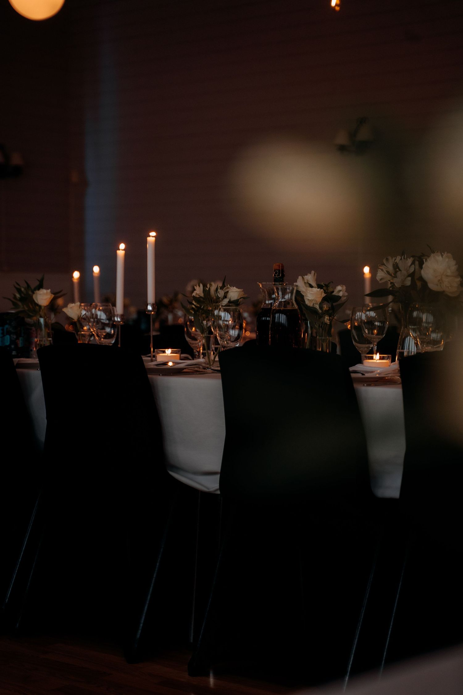 Dimly lit wedding reception table with tall white candles, white floral arrangements, and black chair covers.