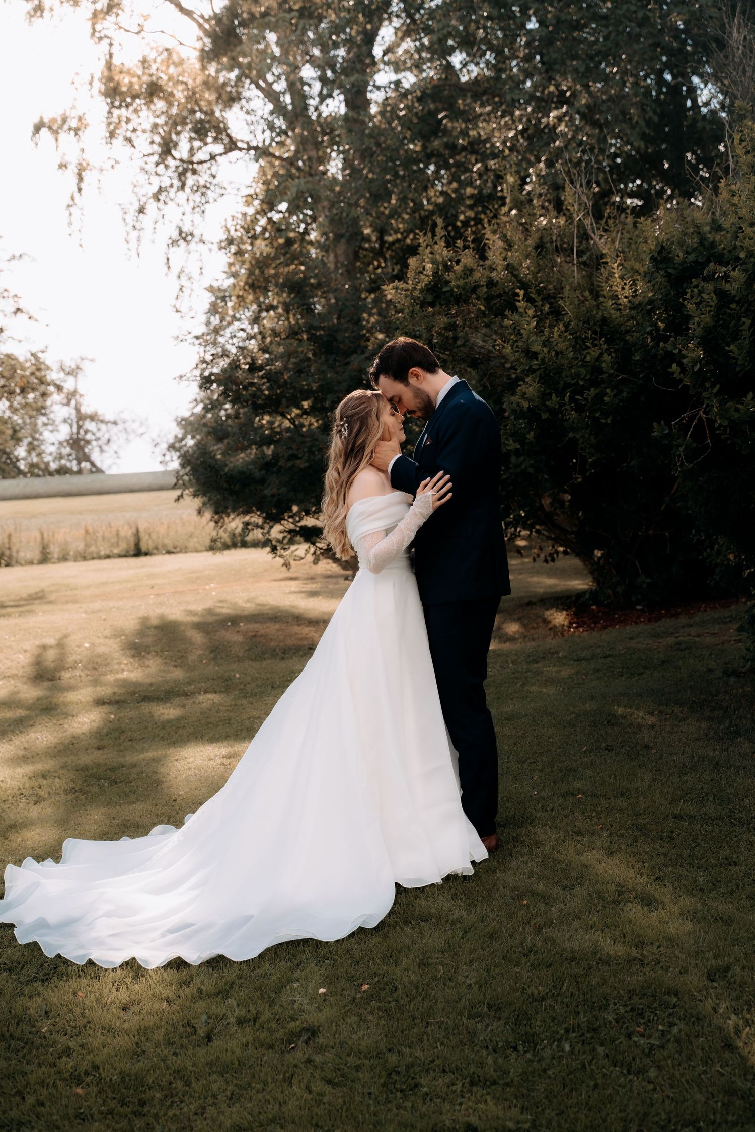 Bride and groom share a romantic forehead touch in a lush garden, bride in a flowing white gown with long train.