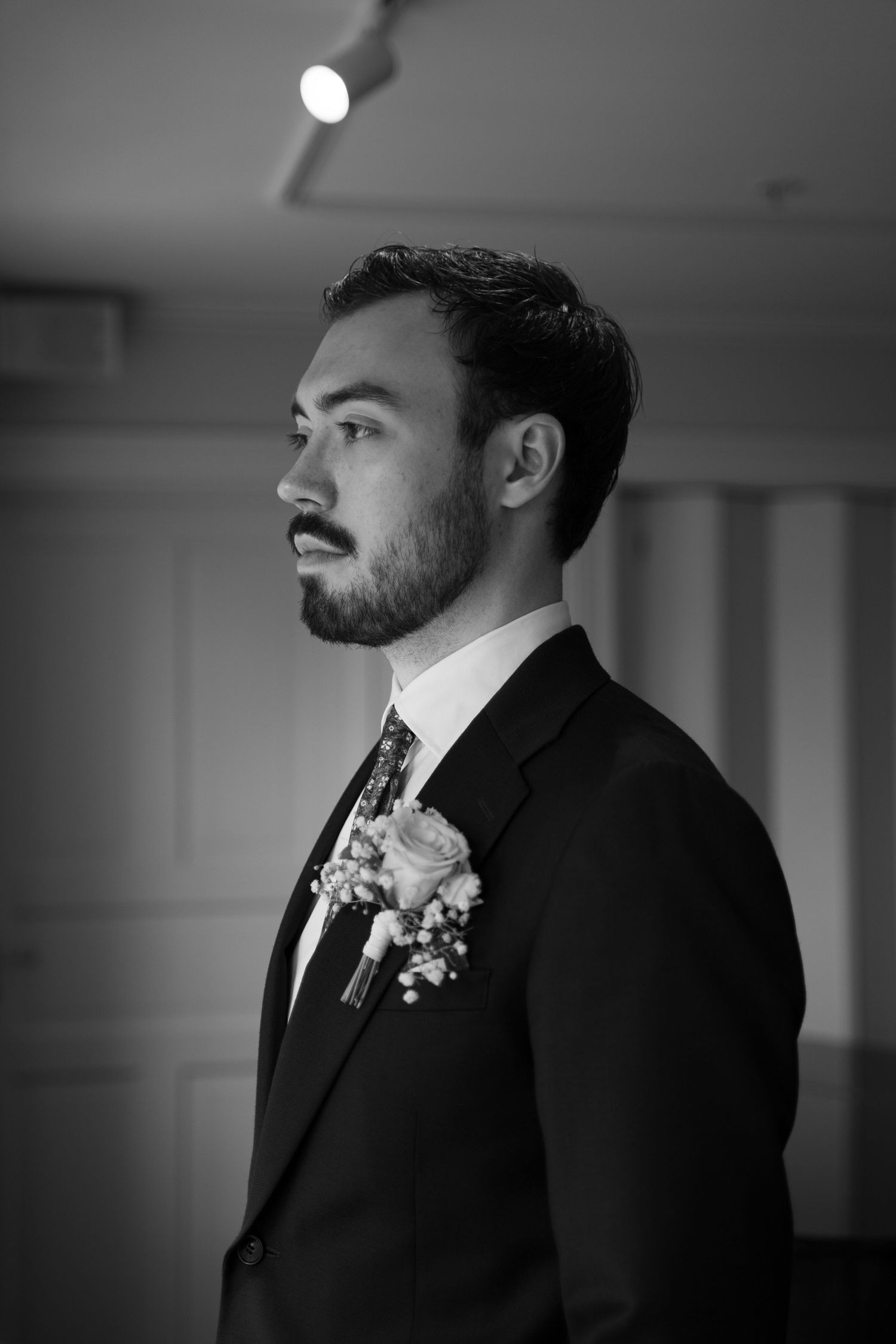 Black and white portrait of bearded groom in dark suit with floral boutonniere gazing thoughtfully to the side.