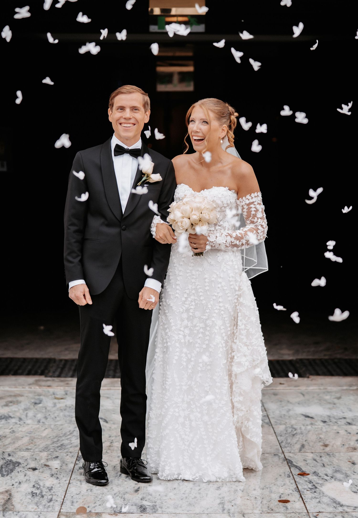 Joyful newlyweds celebrate with white confetti, bride in strapless gown holding bouquet, groom in black tuxedo.
