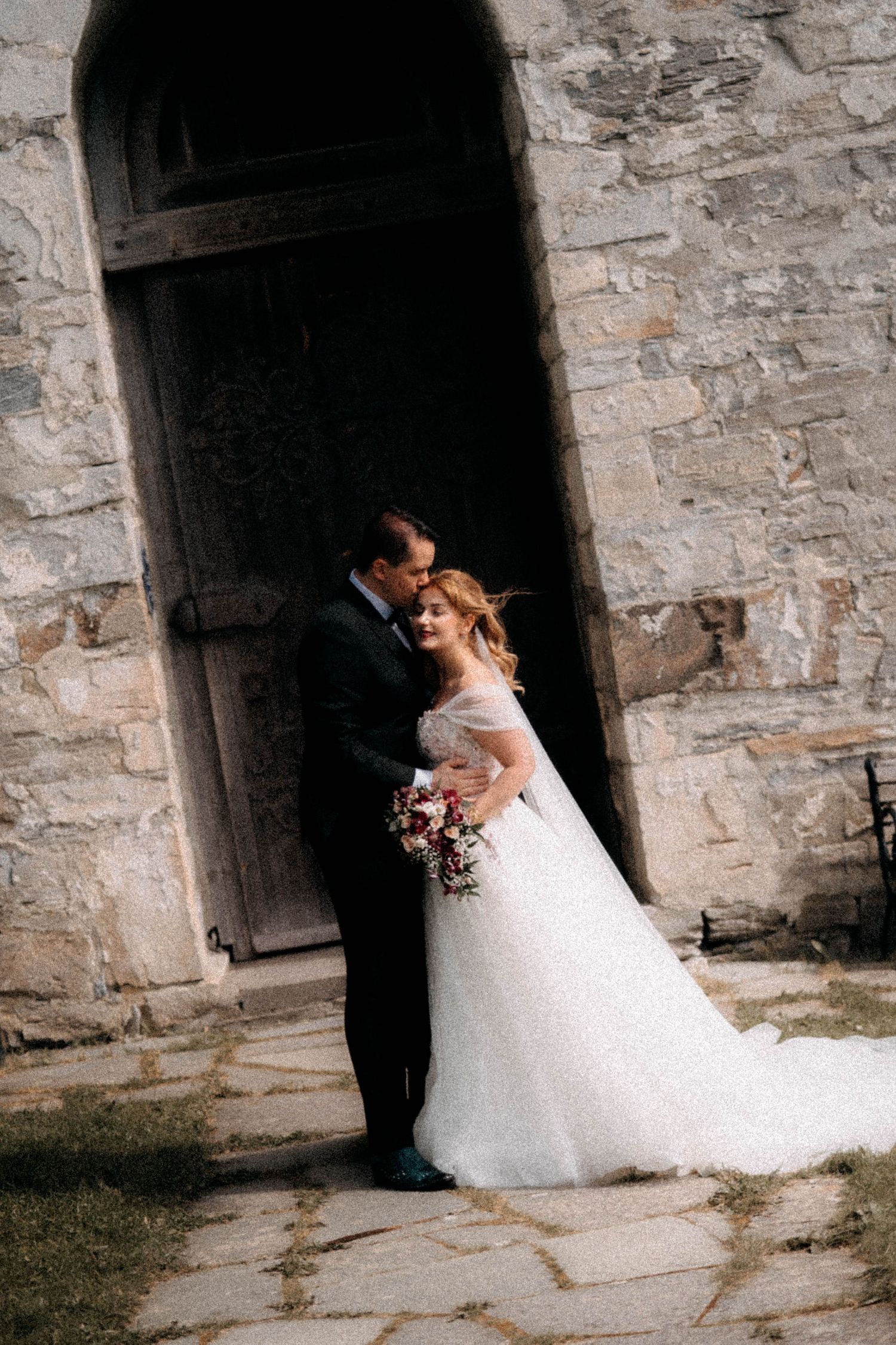 Bride and groom share a romantic embrace outside a rustic stone castle doorway on their wedding day.