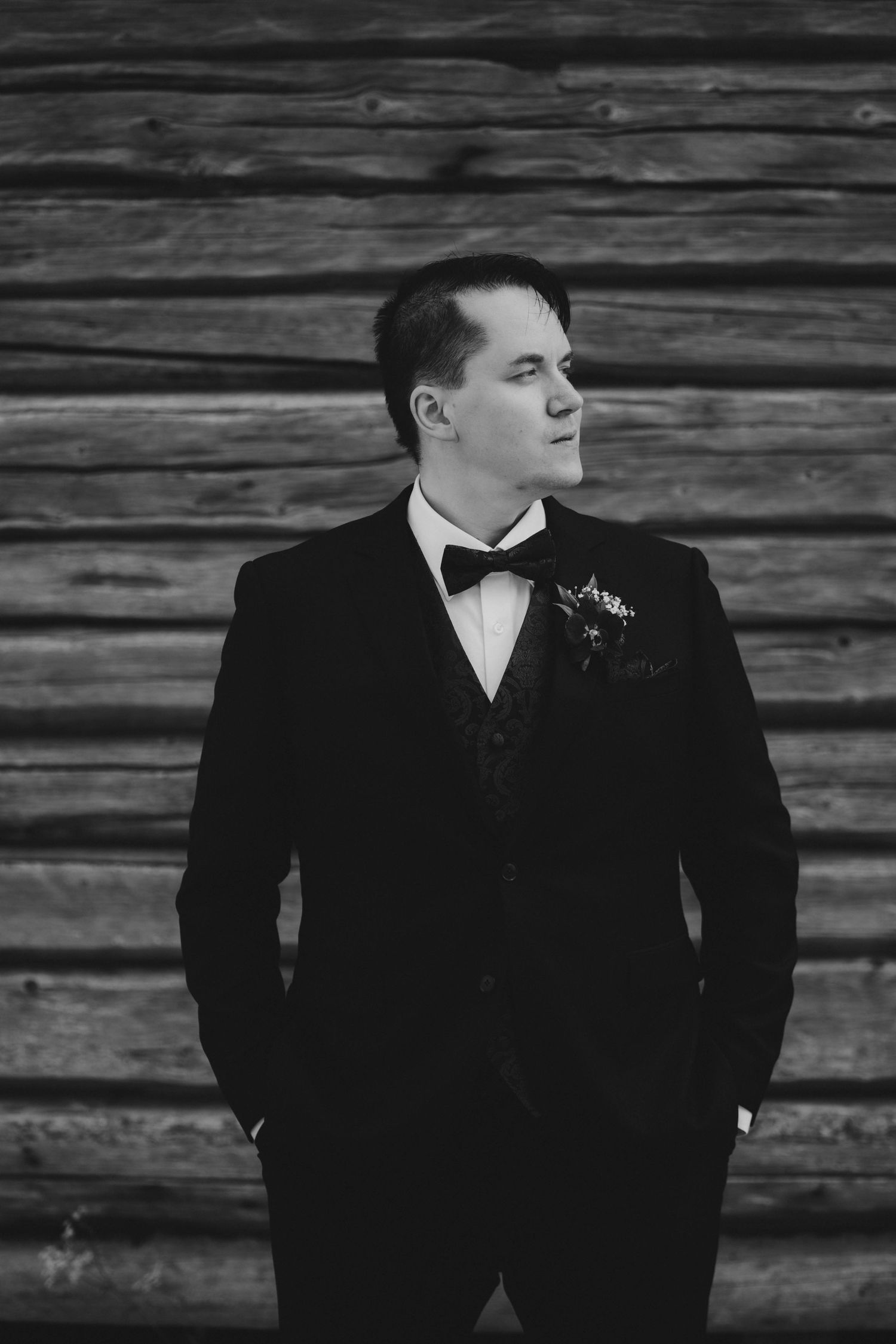 Black and white portrait of a groom in a tuxedo with boutonniere, standing against a rustic wooden wall.