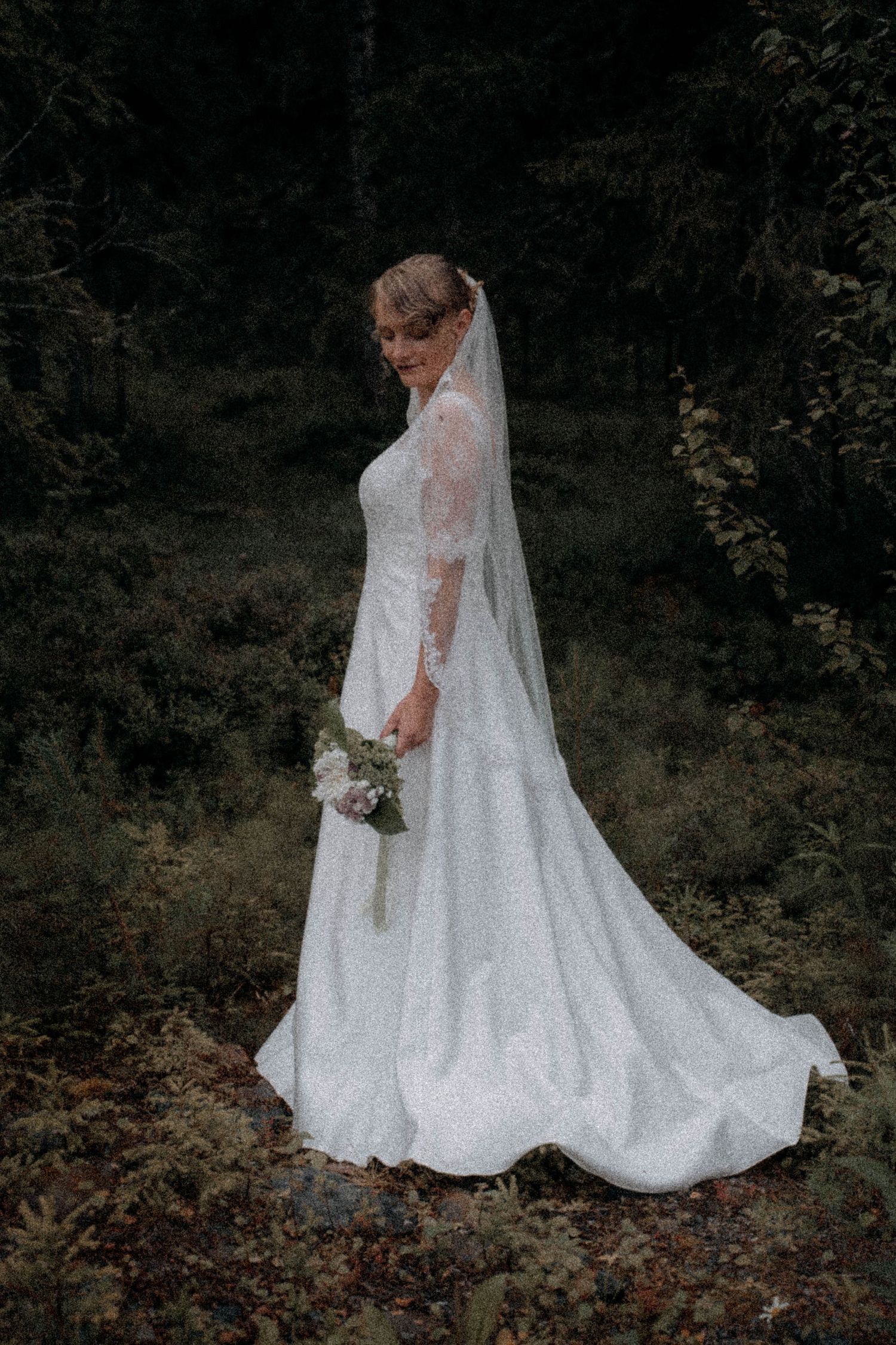 Bride in white lace gown with veil holding bouquet, posing in dark forest setting.