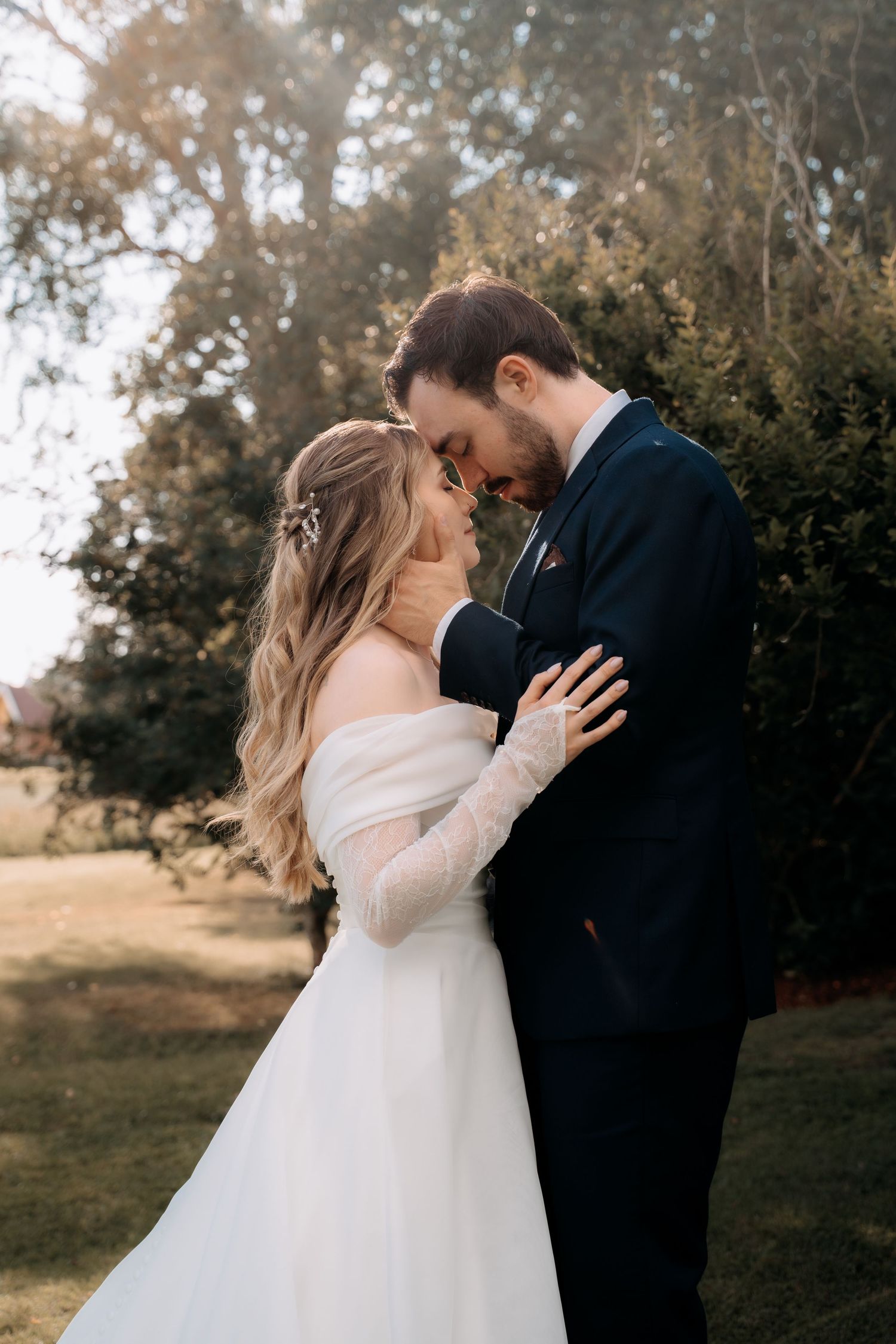 Bride and groom share romantic forehead touch outdoors, with bride in off-shoulder white gown and groom in navy suit.