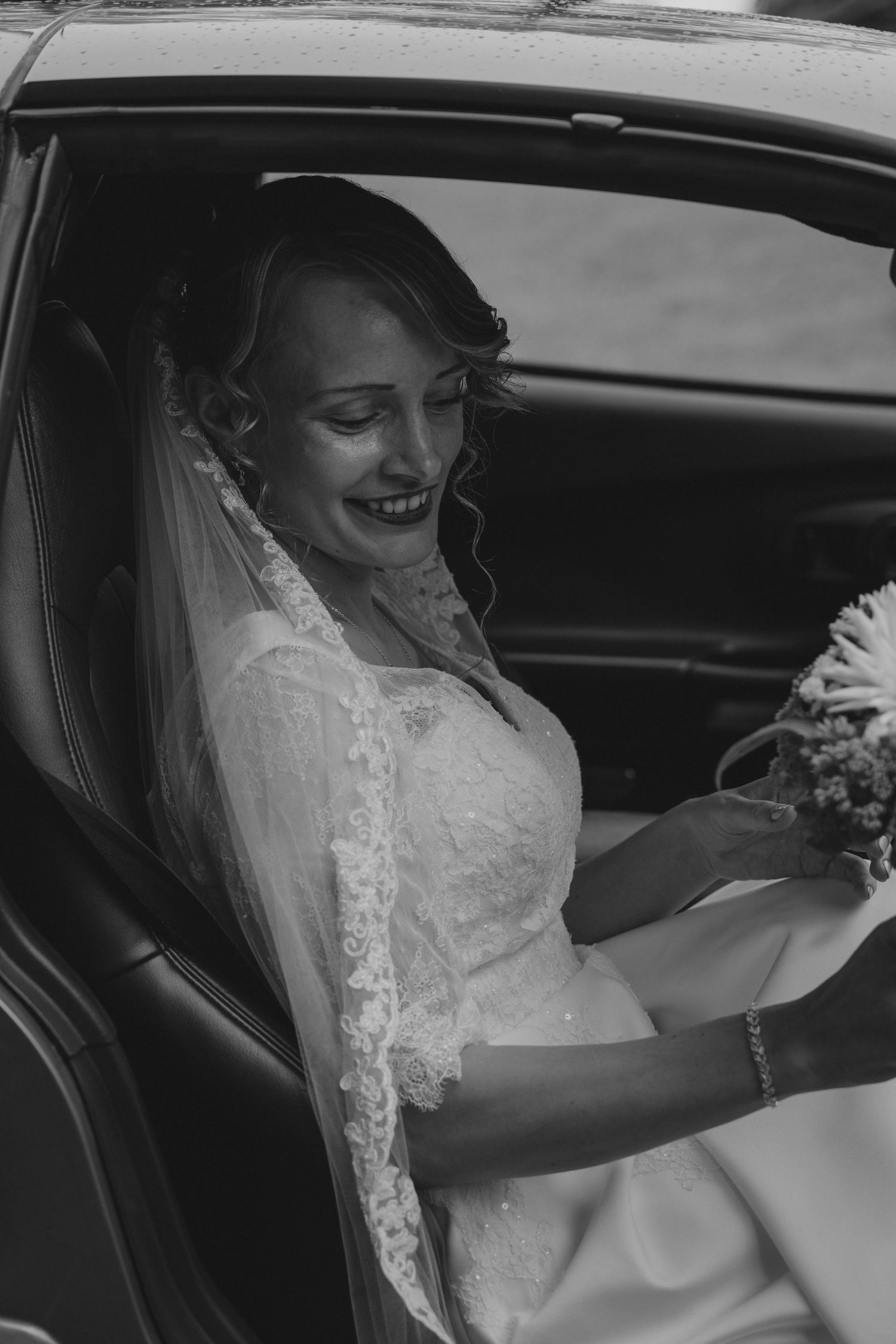 Black and white photo of a smiling bride in lace wedding dress holding bouquet while sitting in a car.