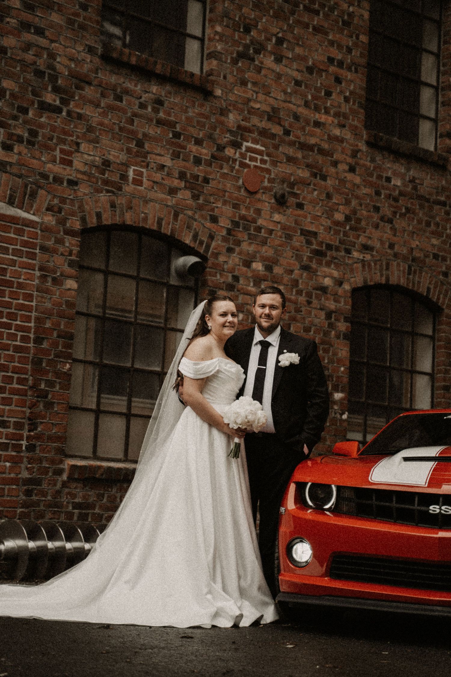Bride and groom posing beside a red Chevrolet Camaro SS in front of a rustic brick building on their wedding day.