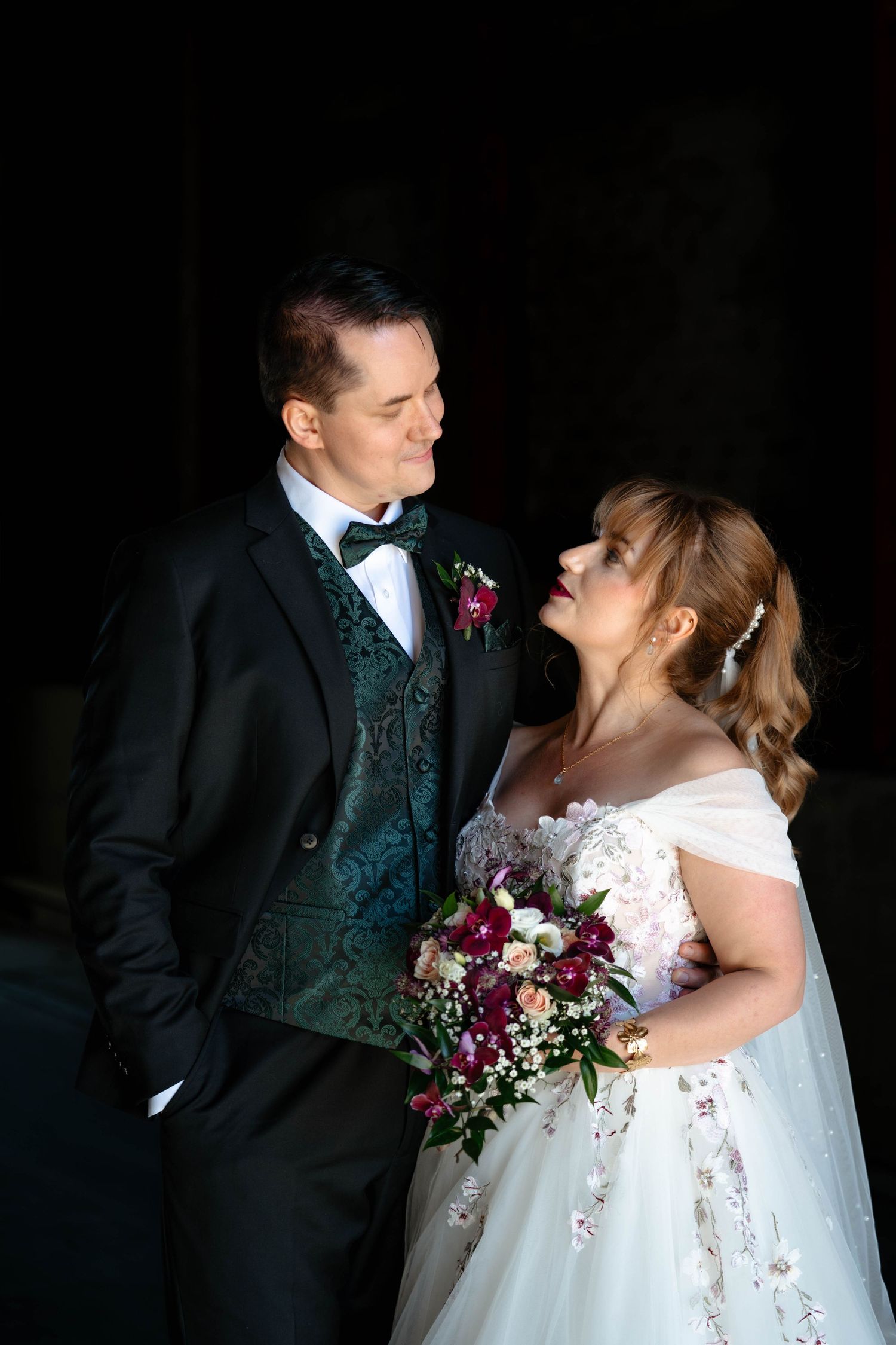 Bride in white gown holding bouquet gazes at groom in black tuxedo with green vest against dark background.