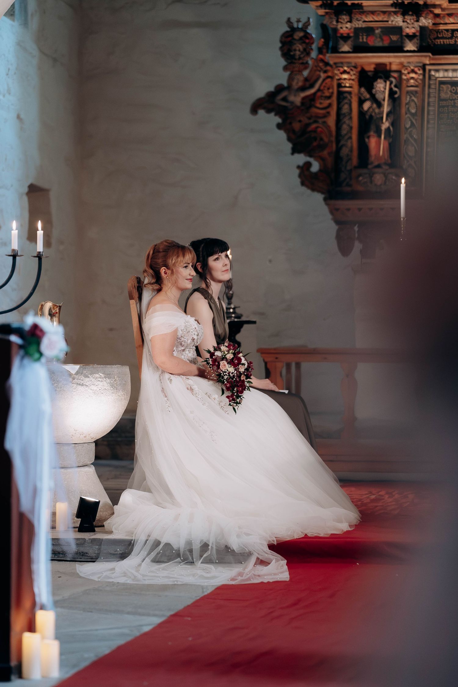 Two brides in white gowns sit together during their wedding ceremony inside a historic church with candlelight.