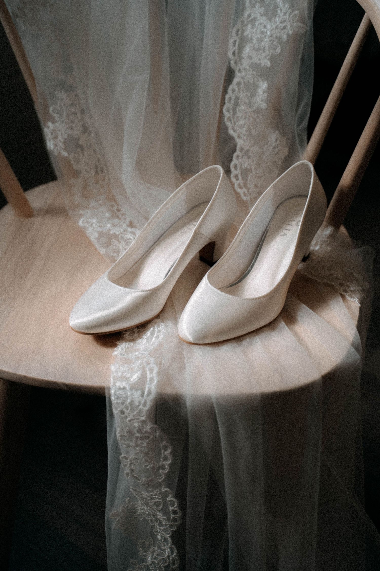 White satin bridal heels displayed on wooden chair with lace wedding veil draped elegantly around them.