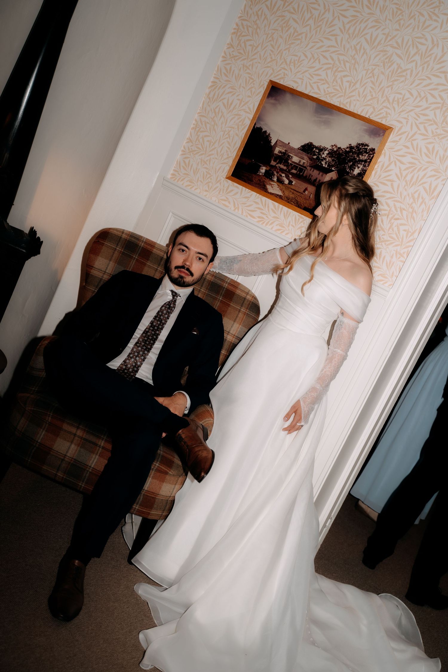 A bride in a white off-shoulder gown stands beside a groom in a dark suit seated in a plaid armchair.