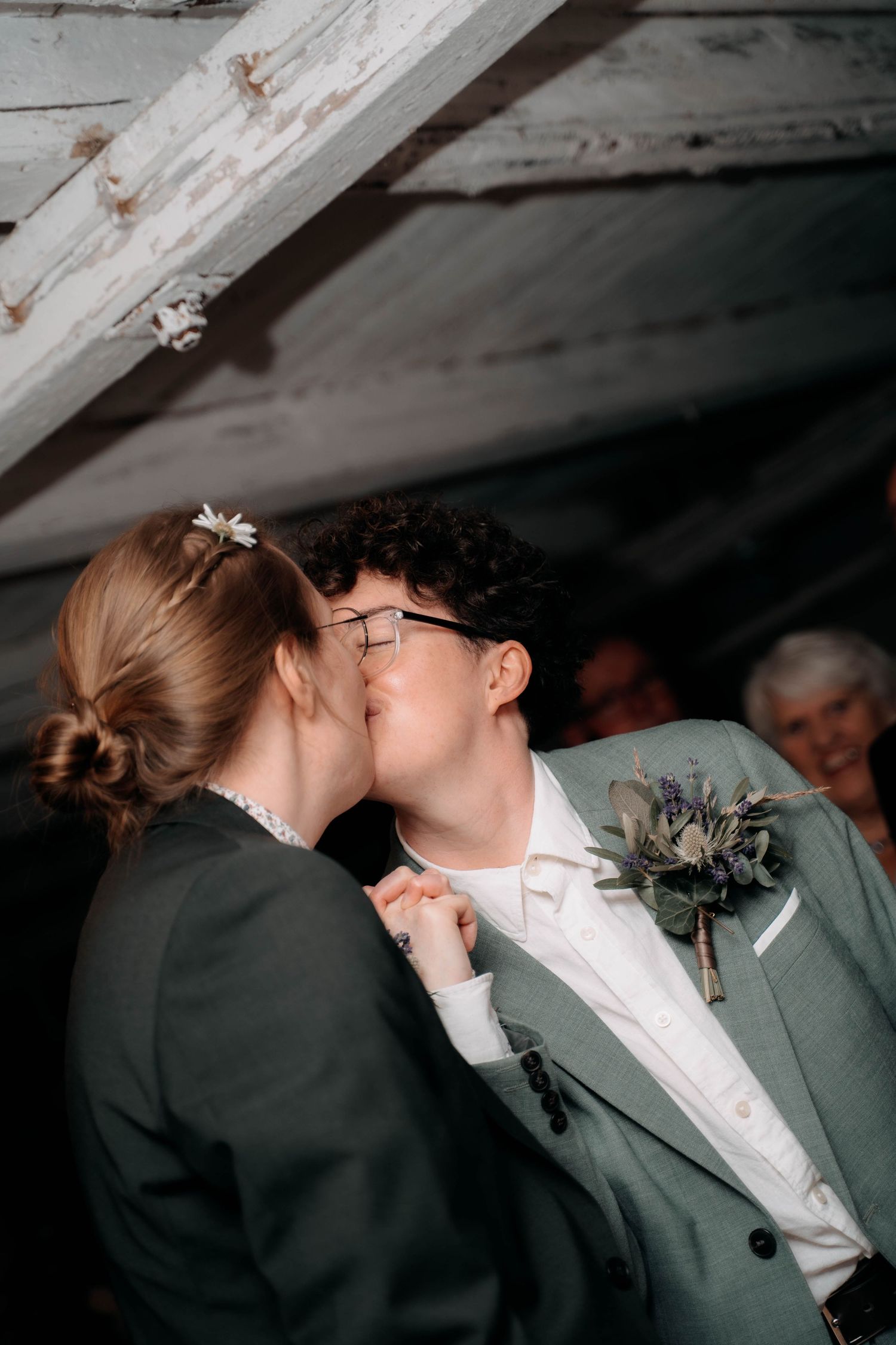 Two brides share a first kiss at their wedding, one wearing glasses with a boutonniere, under rustic wooden beams.