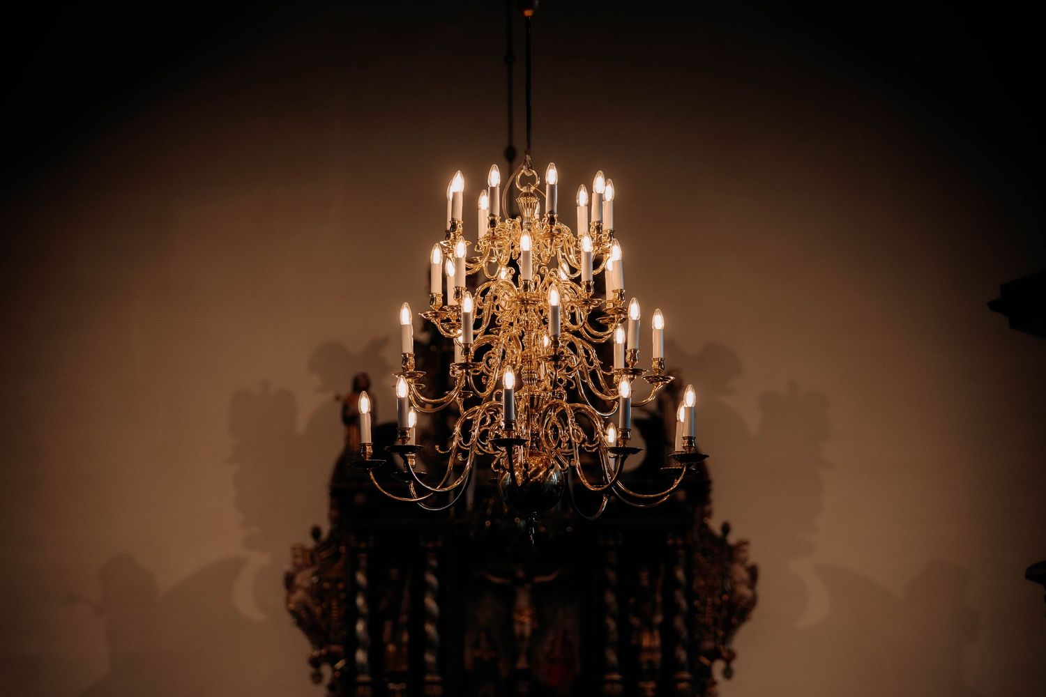 Ornate gold chandelier with lit candles casting warm light and dramatic shadows on a dark wall.