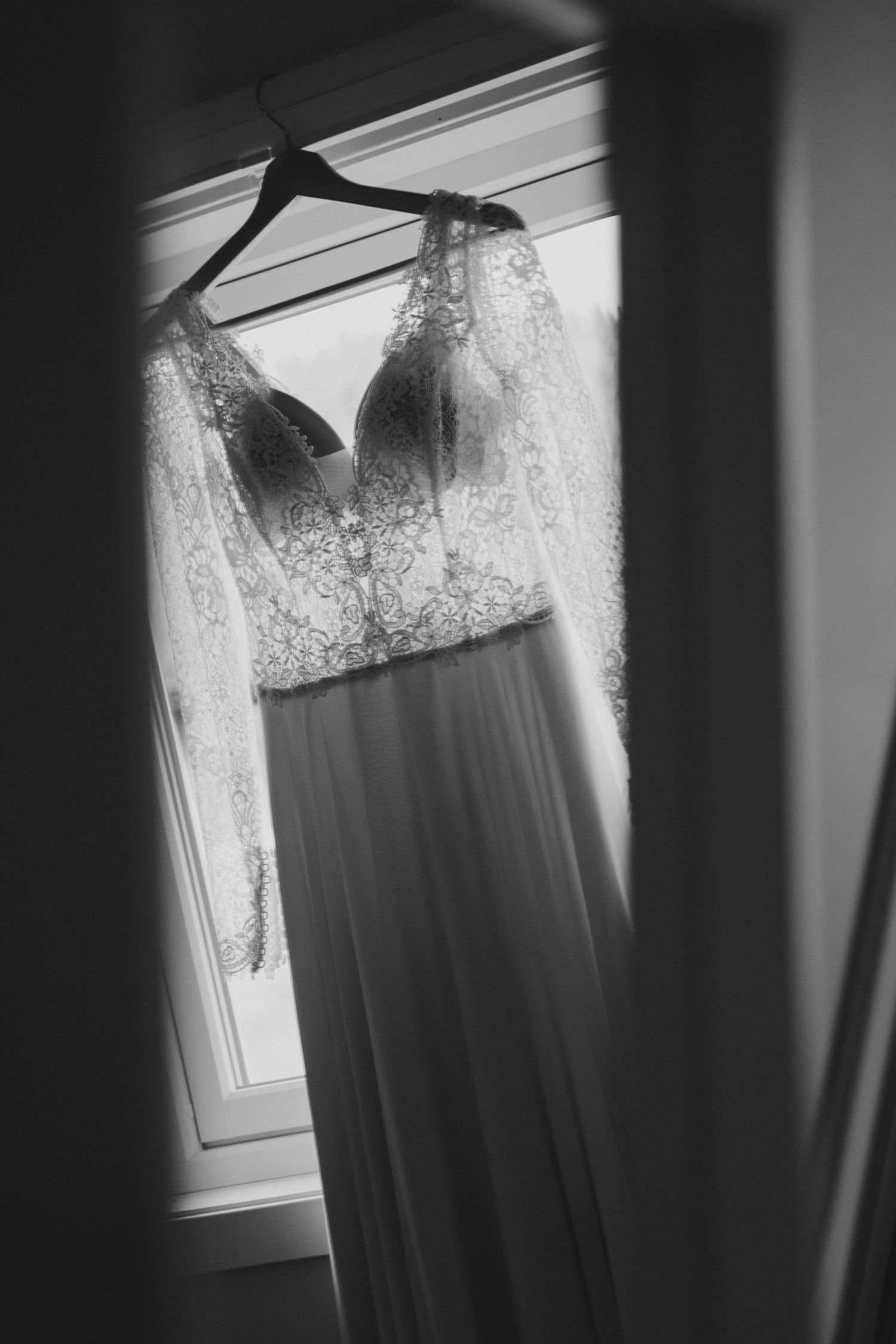 Elegant lace wedding gown with long sleeves hanging in window light, black and white photo.