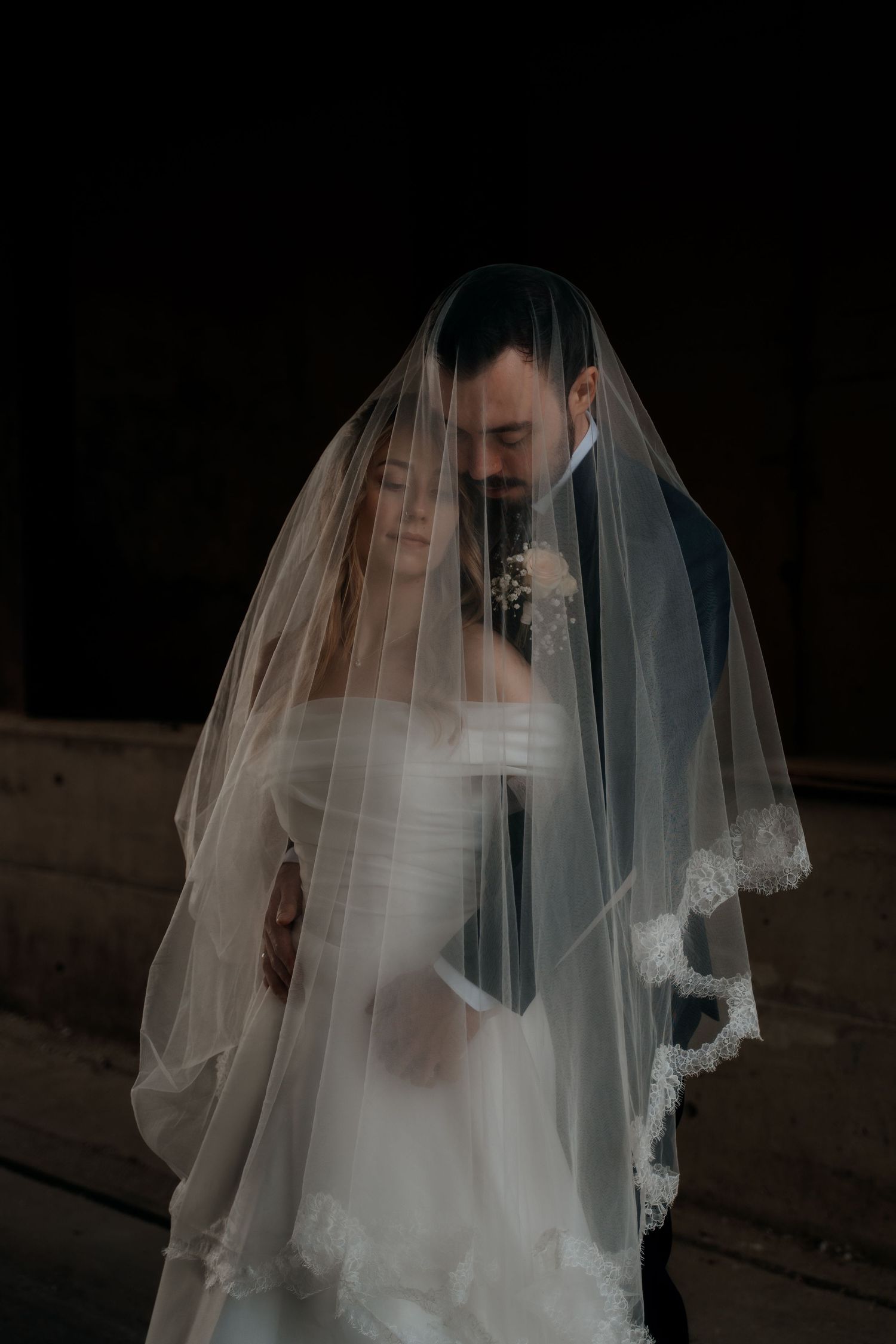 Bride and groom sharing an intimate moment together under a lace-trimmed wedding veil against a dark background.
