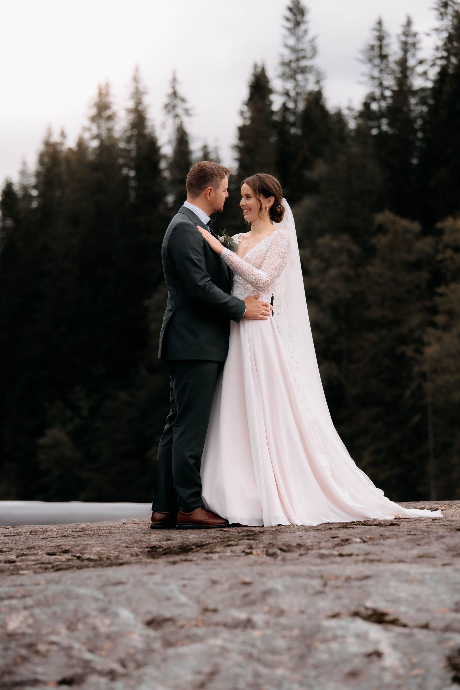 Bride and groom embrace outdoors in front of evergreen forest, wearing elegant wedding attire.