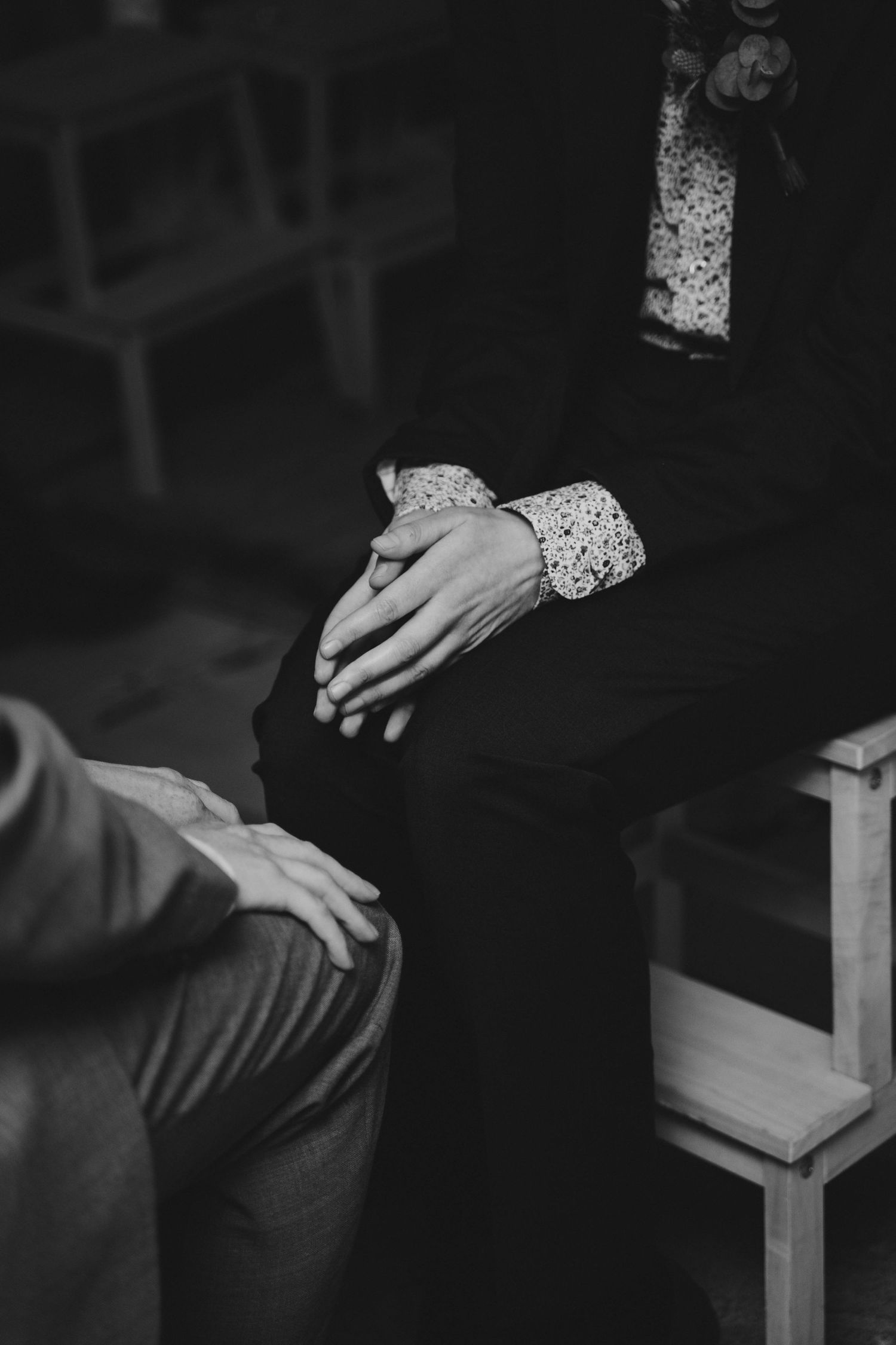 Black and white photo of person sitting with hands clasped wearing patterned blouse cuffs.