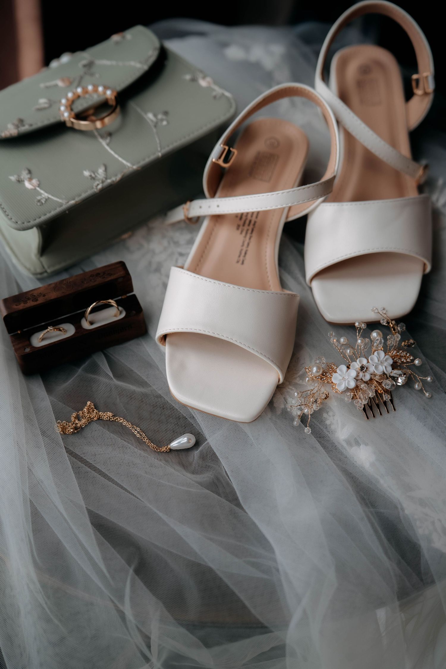 Elegant bridal accessories flat lay featuring white heels, gold hair comb, pearl jewelry, ring box, and sage green clutch on tulle.